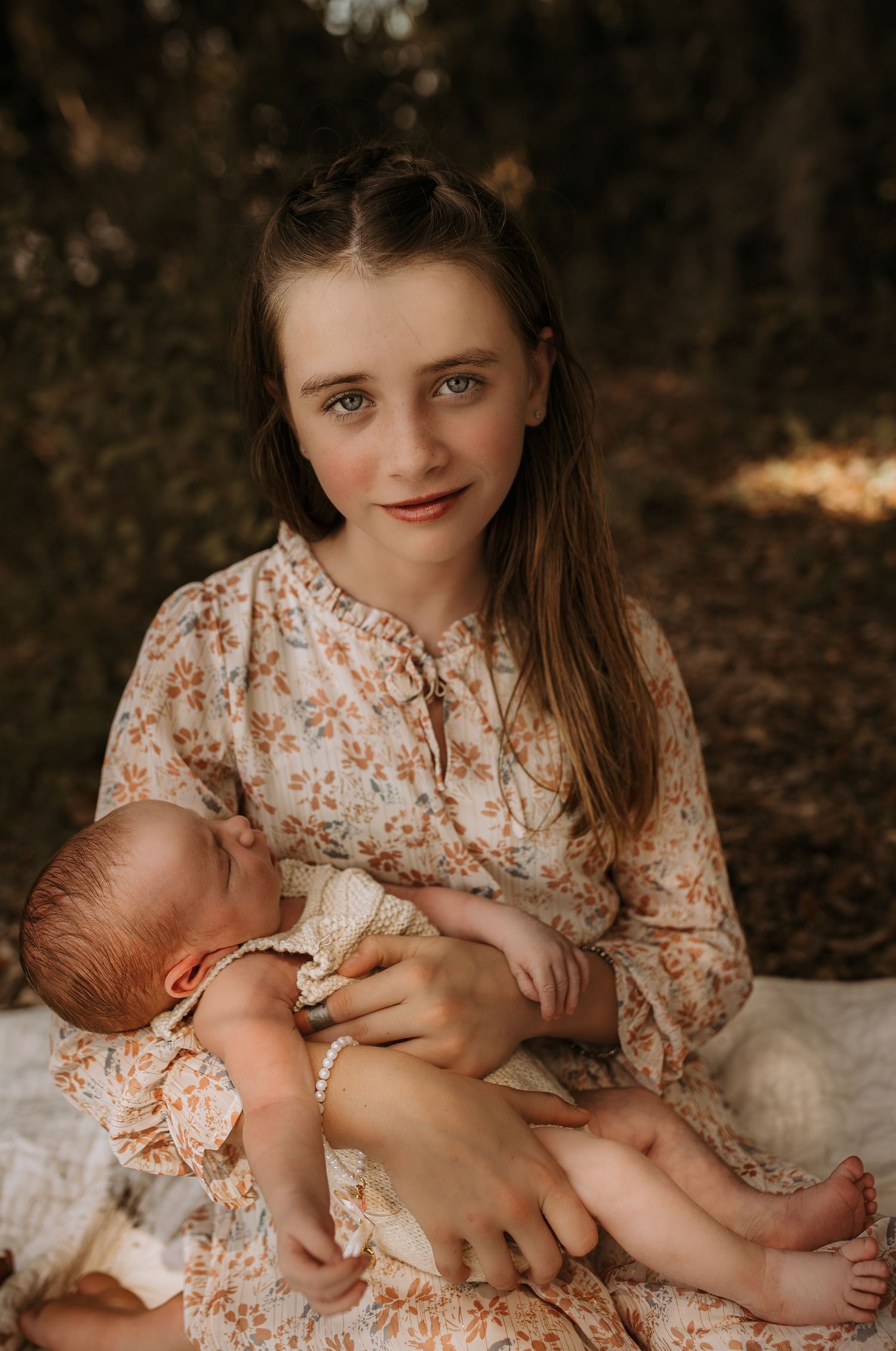 A young girl in a floral dress holds a sleeping newborn baby outdoors in a serene natural setting.