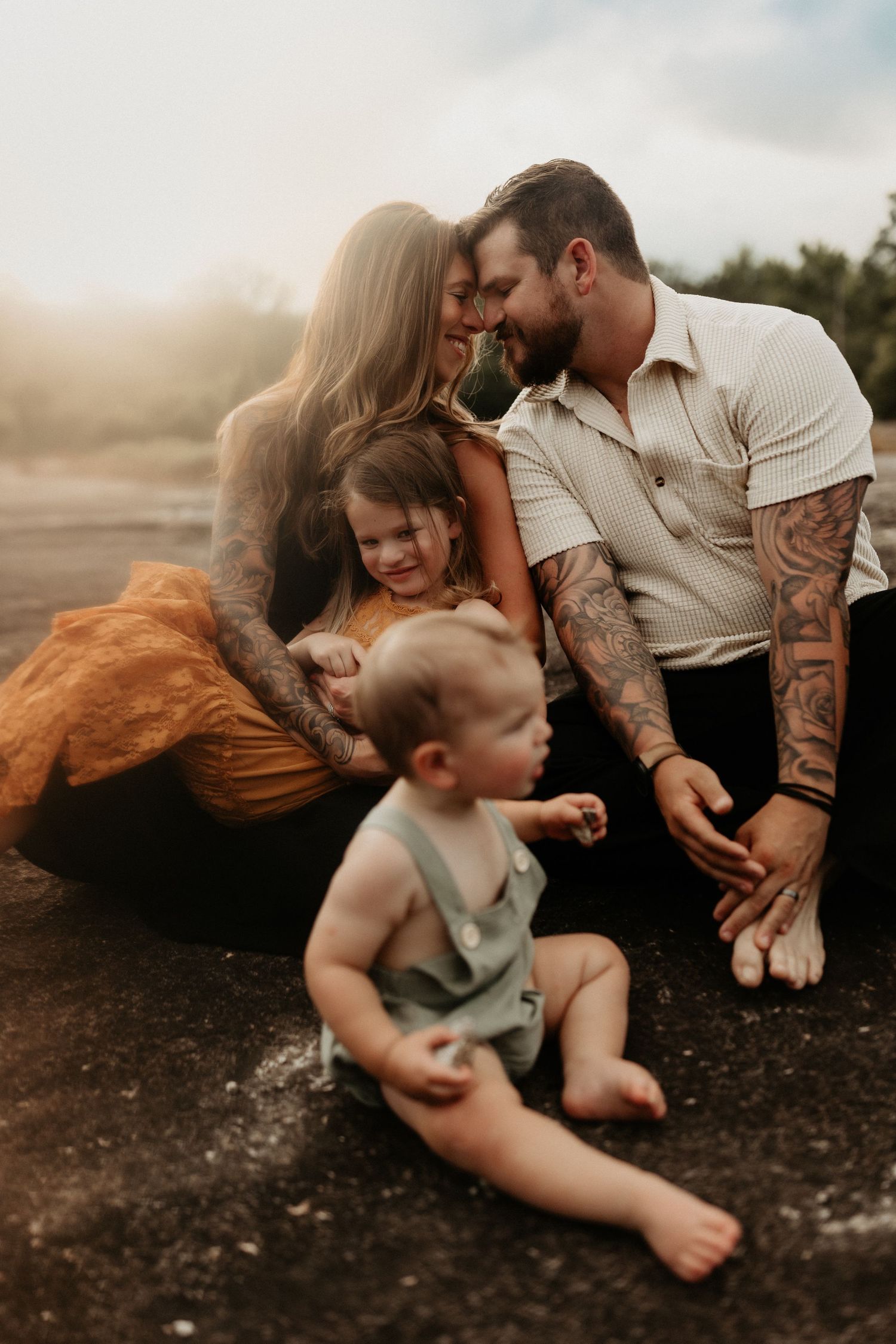 Tattooed parents share a tender moment with their two toddlers during a sun-kissed outdoor family portrait.