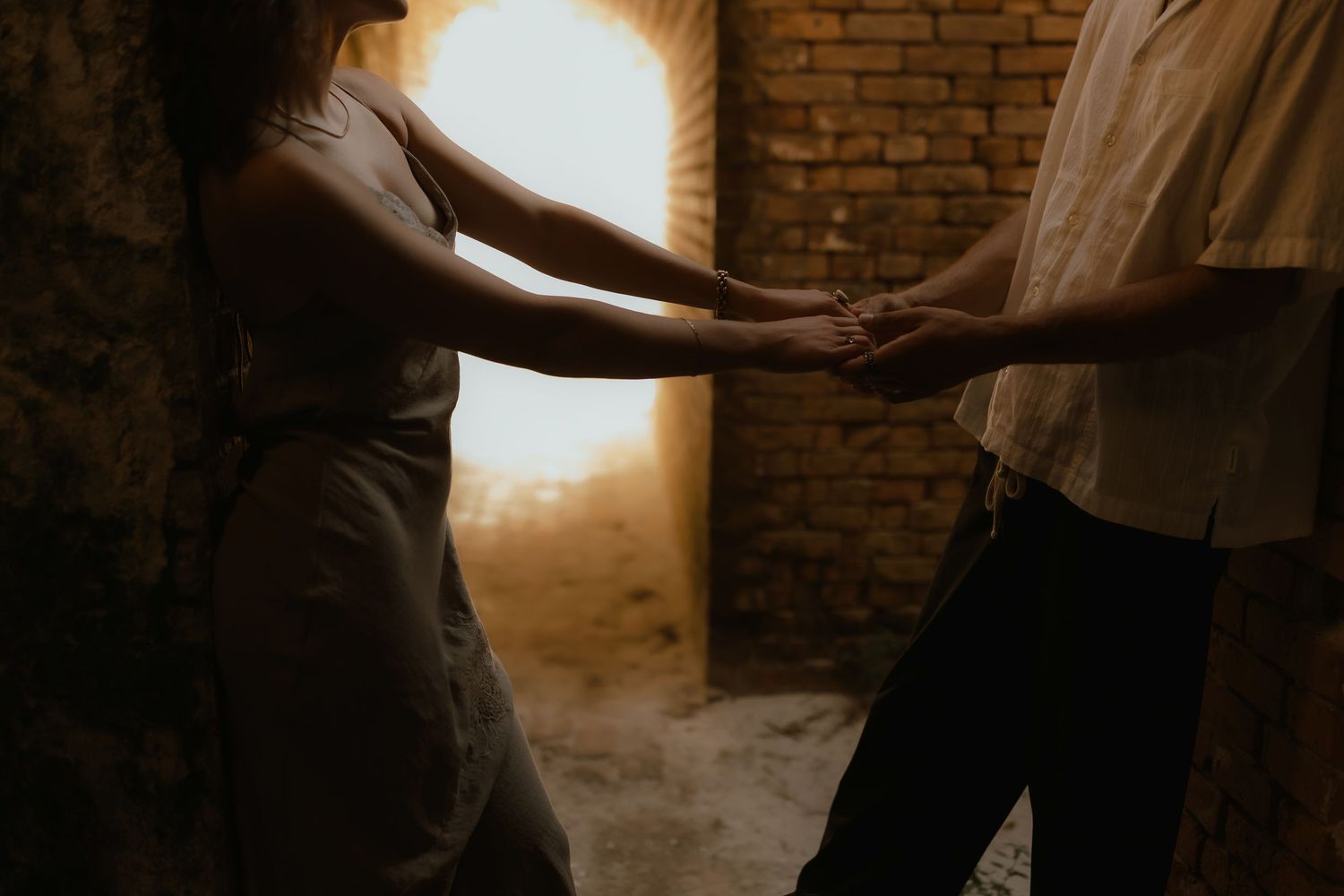 Couple holding hands in a dimly lit rustic room with warm golden light glowing through an arched doorway.