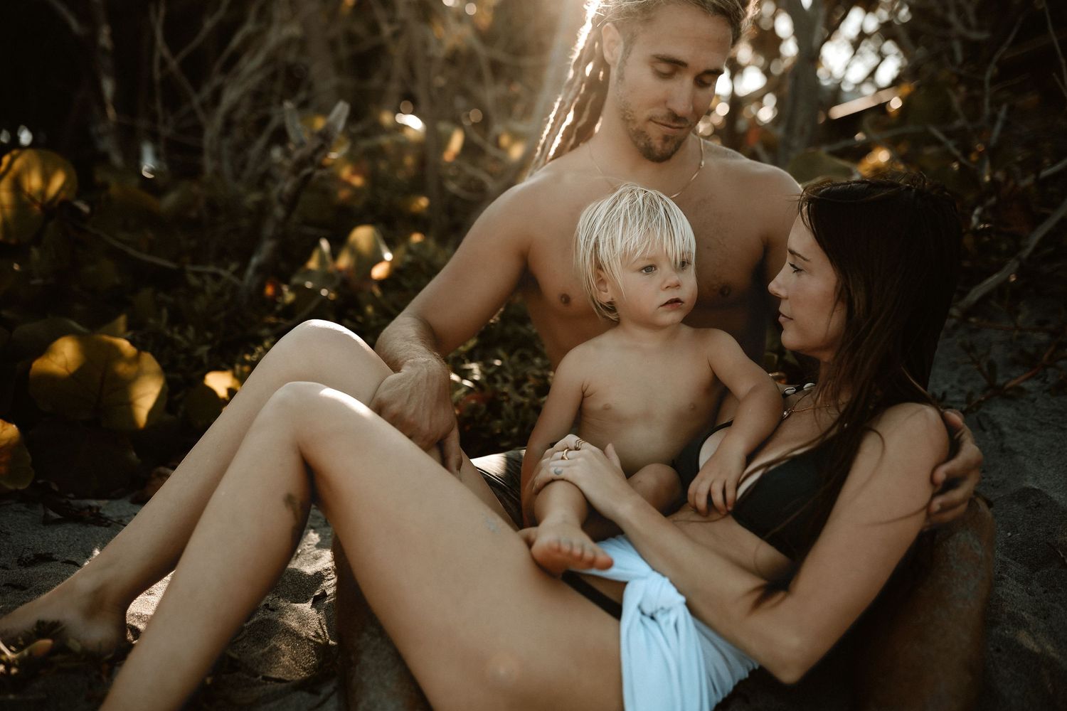 Family of three enjoying a golden hour beach moment, parents relaxing with their young blonde toddler in a natural setting.