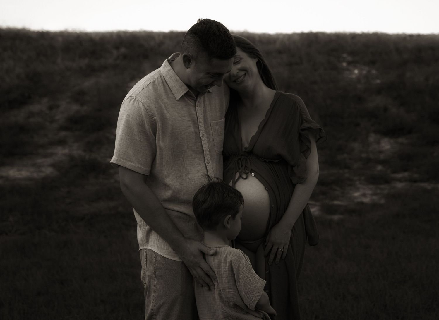 Black and white maternity photo of pregnant woman, partner, and child embracing outdoors at dusk.