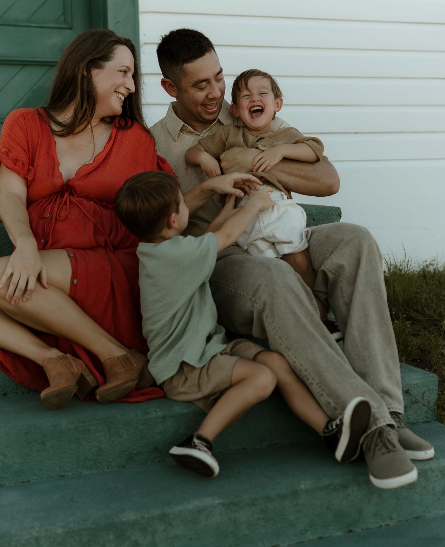 Happy family sitting on porch steps, pregnant mom in red dress, dad holding laughing toddler, young boy joining in.