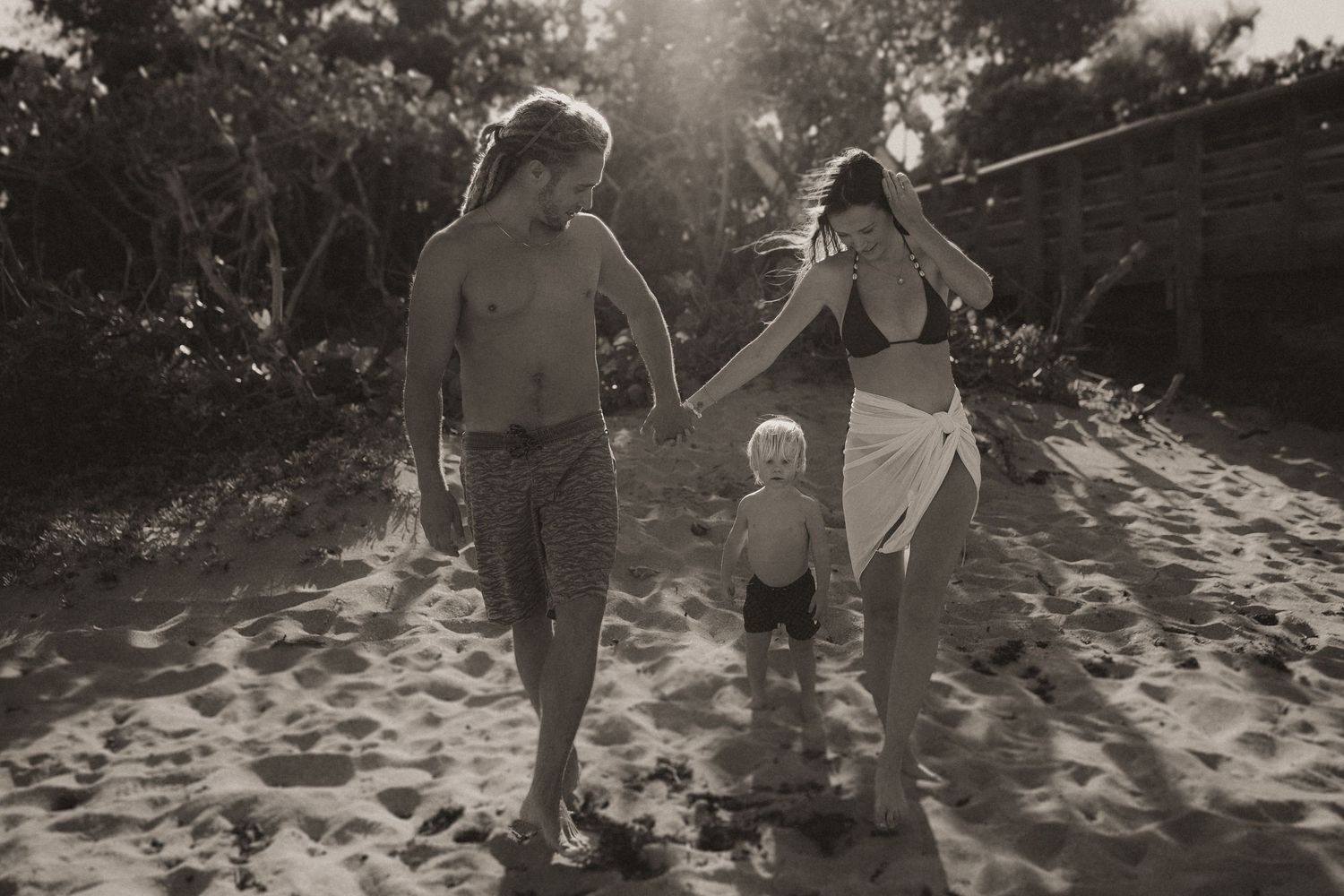Black and white photo of a family with a toddler walking on a sandy beach surrounded by lush trees.