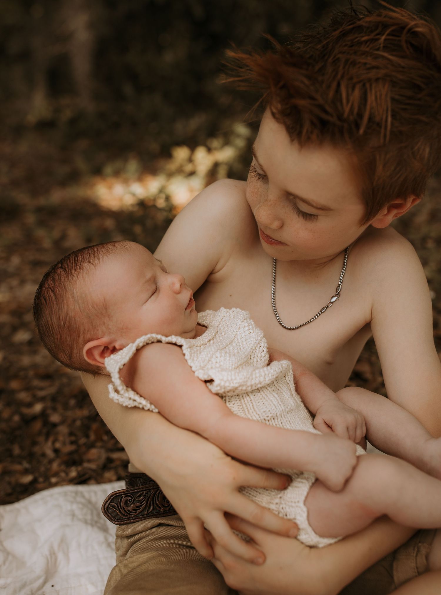 A red-haired boy tenderly holds a sleeping newborn baby in his arms while seated outdoors.