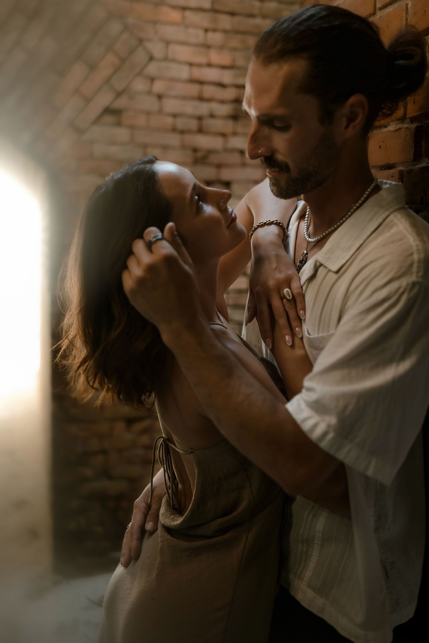 Romantic couple embracing tenderly against a rustic brick wall in warm, moody lighting.