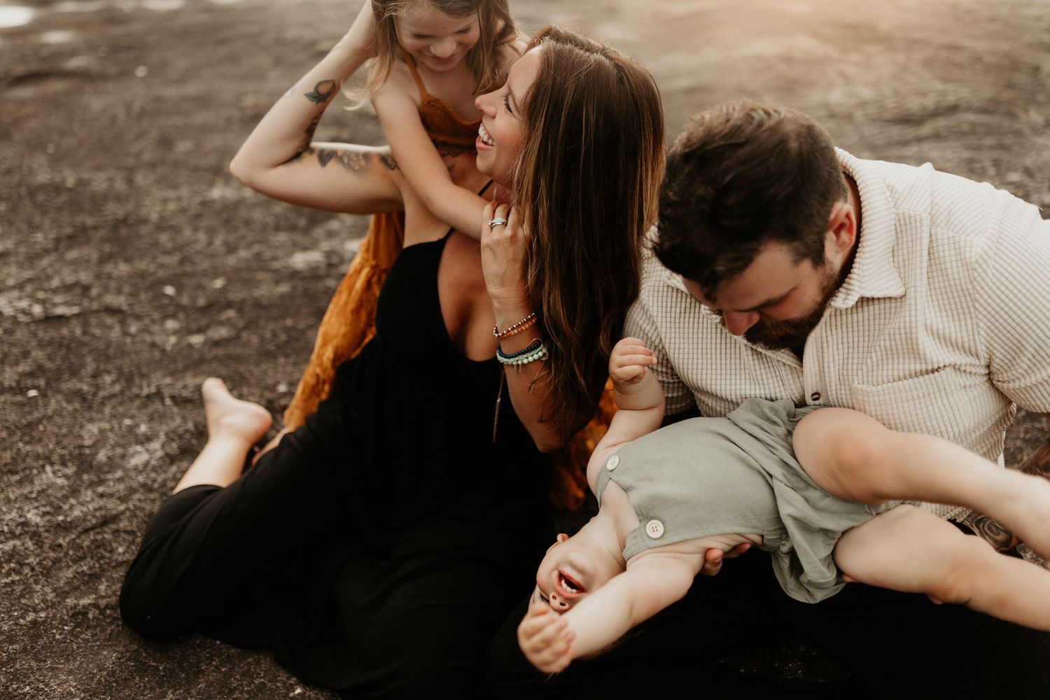 Family laughing and playing together on the ground outdoors during a relaxed lifestyle photo session.