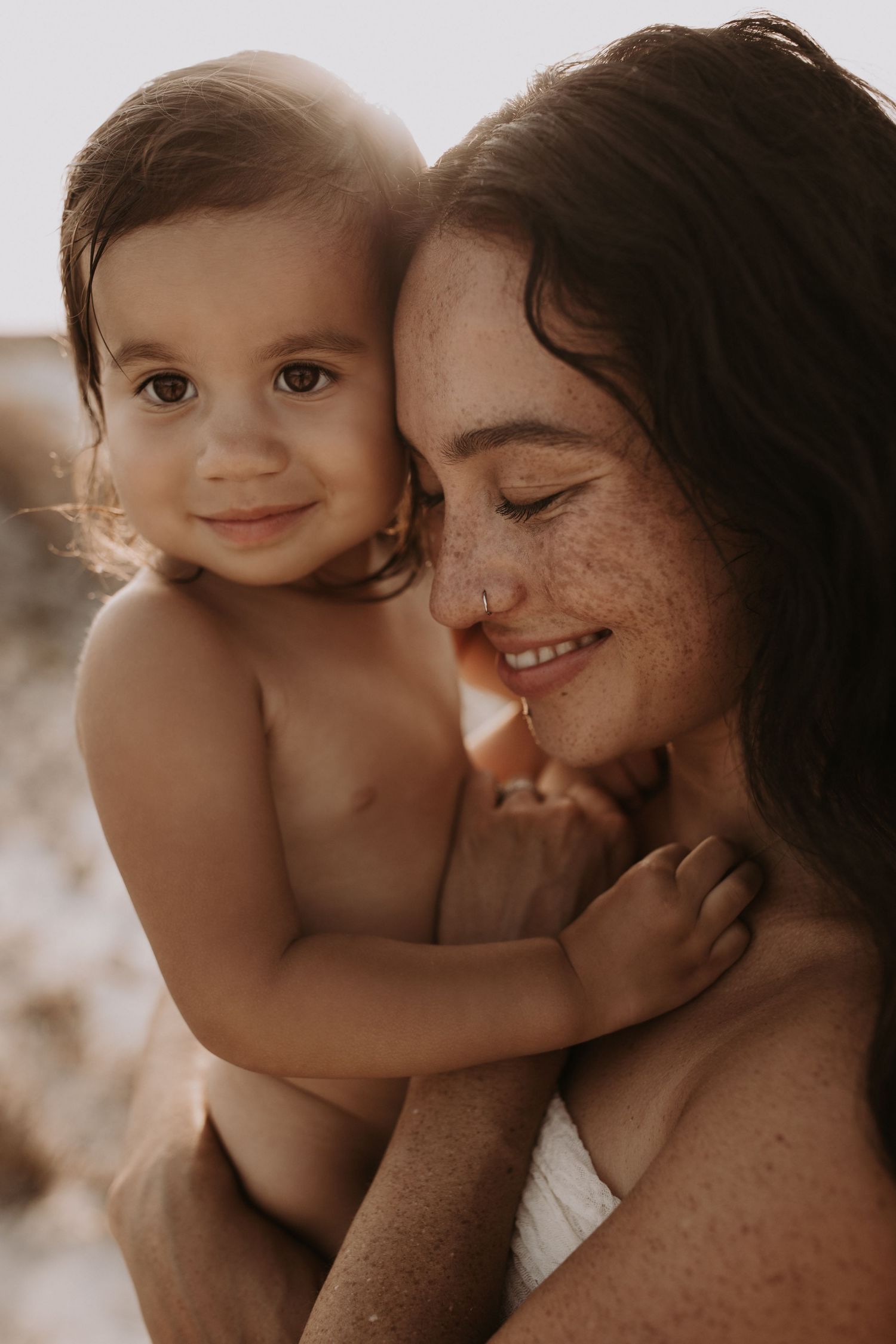 Smiling freckled mother cuddling happy toddler at beach in soft golden light.
