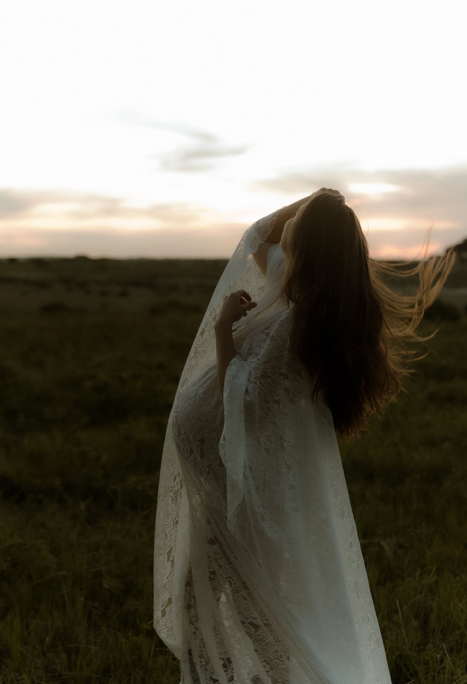 A bride in a flowing white lace gown and veil stands in an open field at sunset with her hair blowing in the wind.