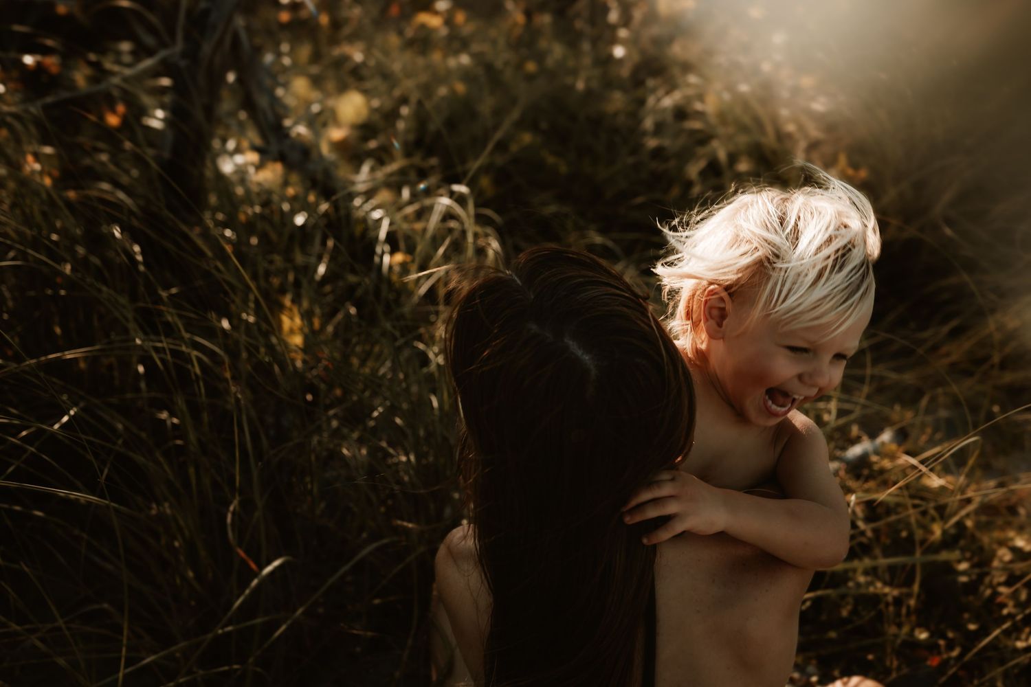 Young blonde child hugging a dark-haired woman in a sunlit natural setting.