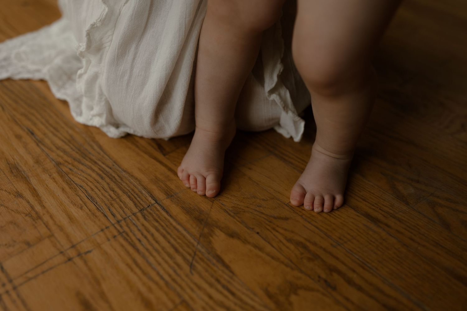 Close-up of chubby baby feet standing on hardwood floor beside a white dress hem.