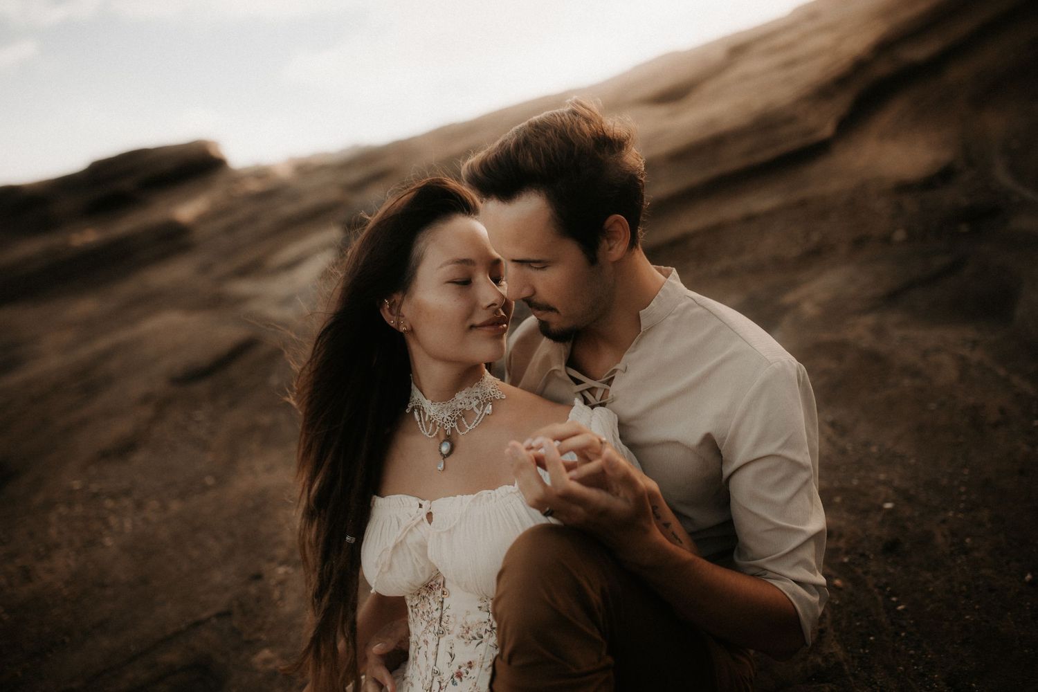 Romantic couple embracing on rocky terrain at sunset, woman in white off-shoulder dress with delicate necklace.
