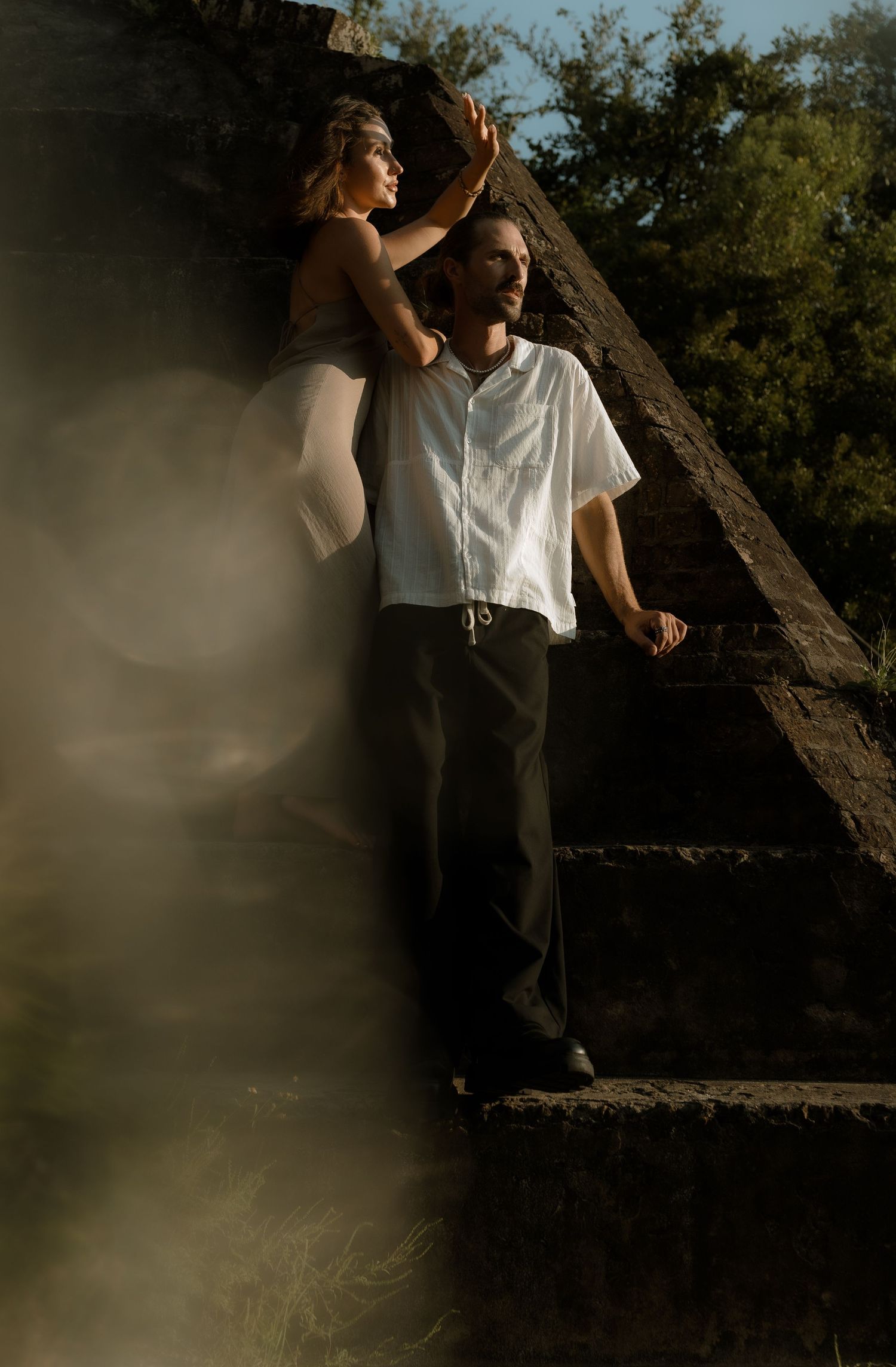 A couple poses dramatically on stone steps, man in white shirt and dark pants, woman in white dress, surrounded by misty light.