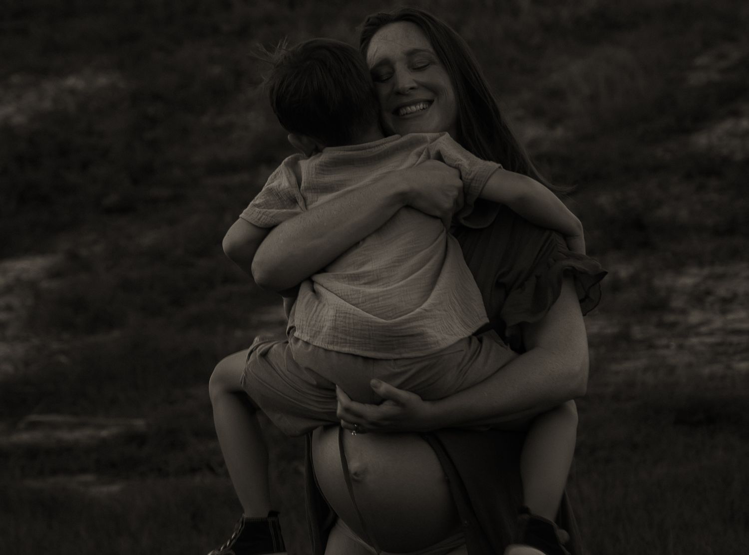Black and white photo of a laughing woman hugging a child outdoors in a grassy field.