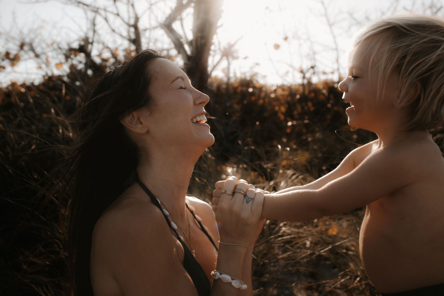 A joyful mother and young child share a tender moment outdoors, laughing together in warm golden sunlight.