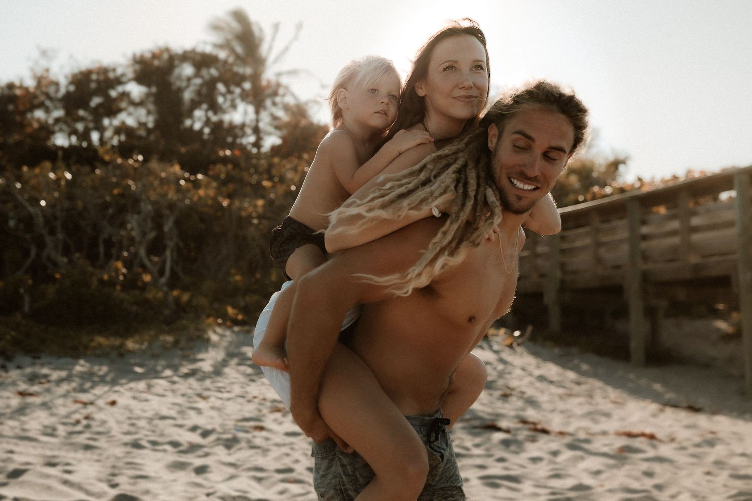 Joyful man giving piggyback ride to woman and child on sunny beach with golden light.