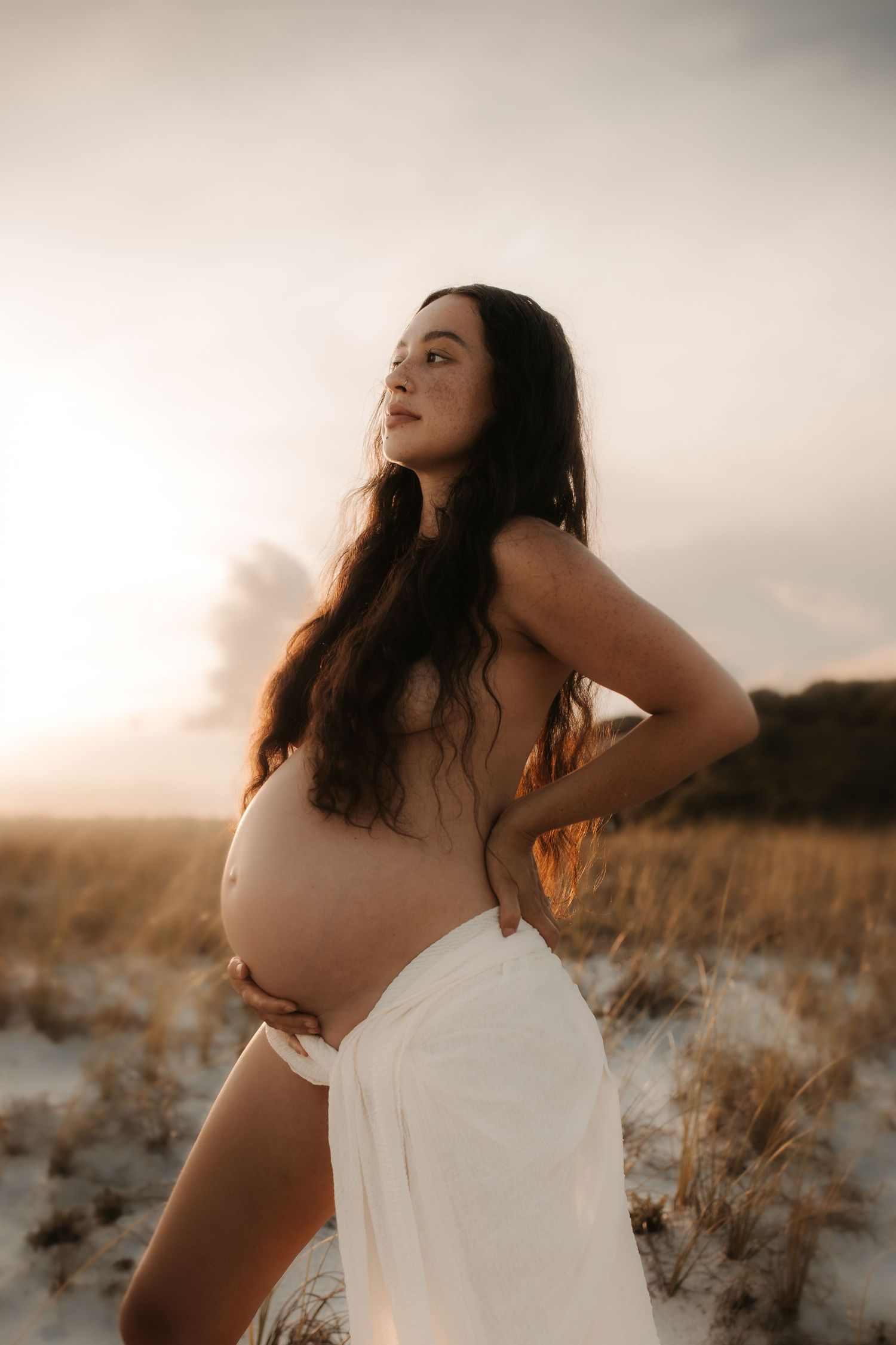 Pregnant woman with long dark hair poses on sandy dunes at sunset wrapped in white fabric.