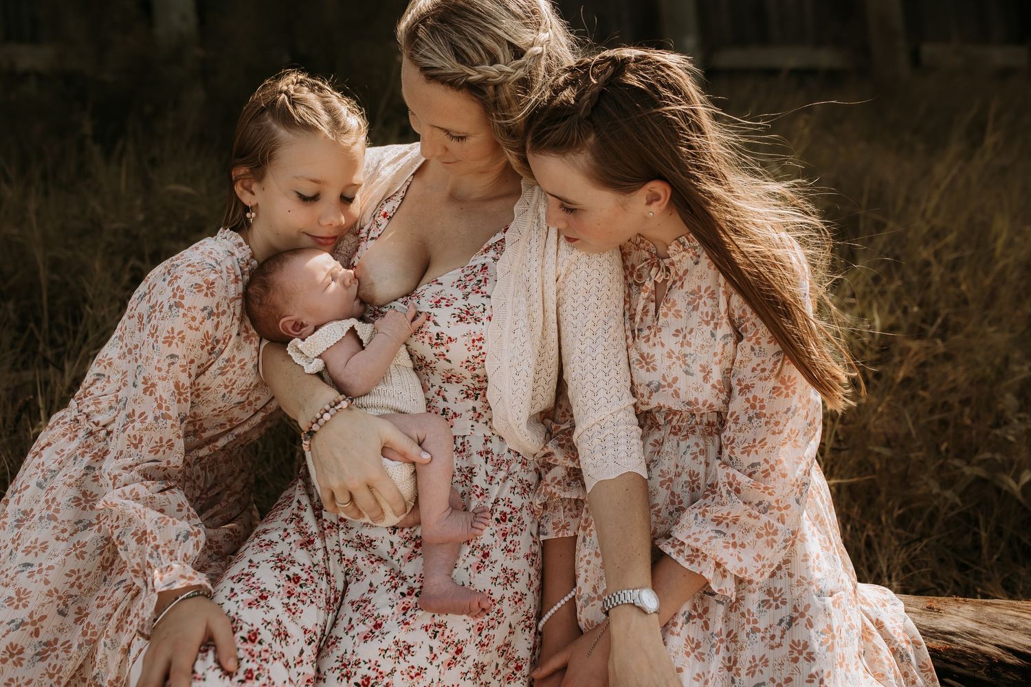 A mother in a floral dress holds her sleeping newborn while two daughters lean in close outdoors.