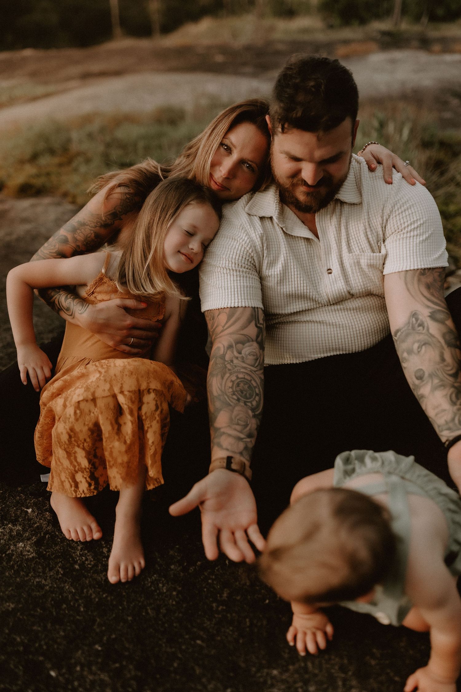 Tattooed couple sitting outdoors with two young children in a warm, candid family portrait at golden hour.