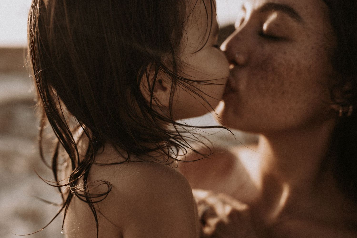 Freckled mother gently kissing young child in warm golden sunlight outdoors.
