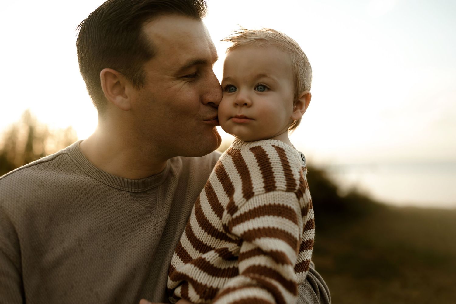 A father tenderly kisses his toddler outdoors at sunset, child wearing a brown striped sweater.