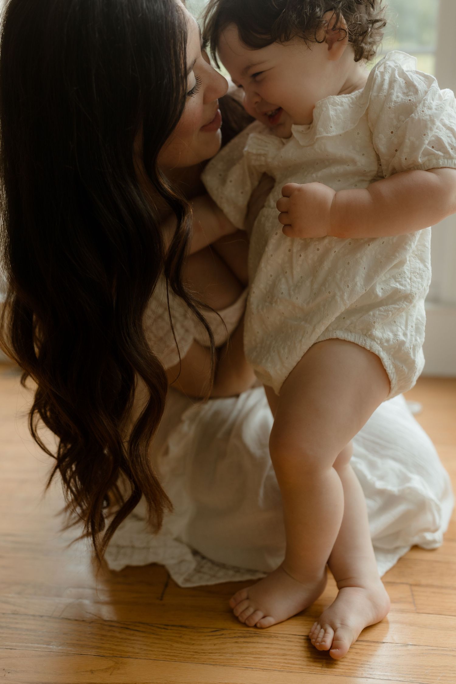 A mother with long dark hair holds her laughing baby in a white romper, sharing a tender moment on a warm wooden floor.