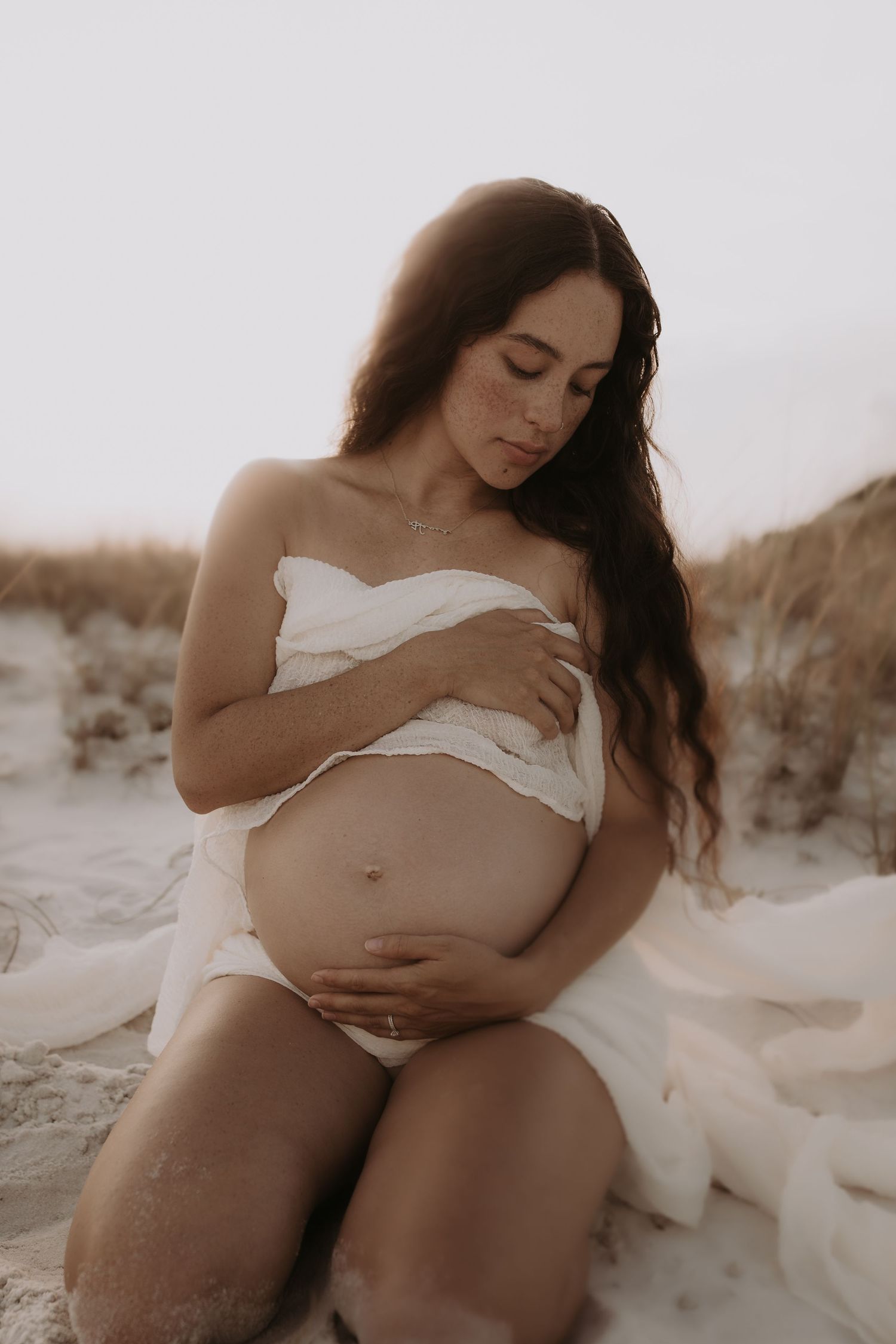 Pregnant woman in white wrap sitting on sandy beach, cradling her baby bump in a serene maternity photo.
