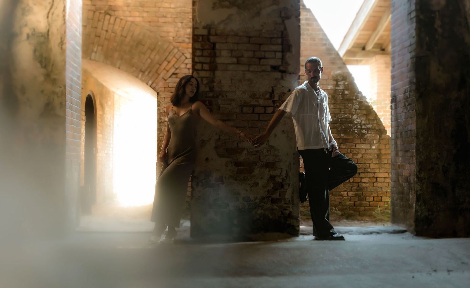 Two people stand in a rustic brick building with dramatic light streaming through windows.