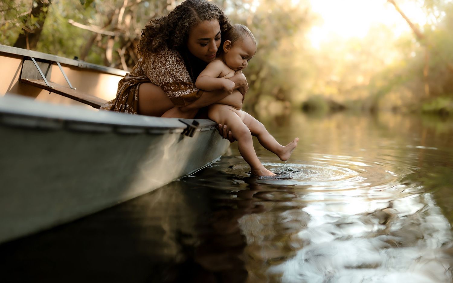 A mother and baby sit on the edge of a canoe, gently touching the calm river water together.