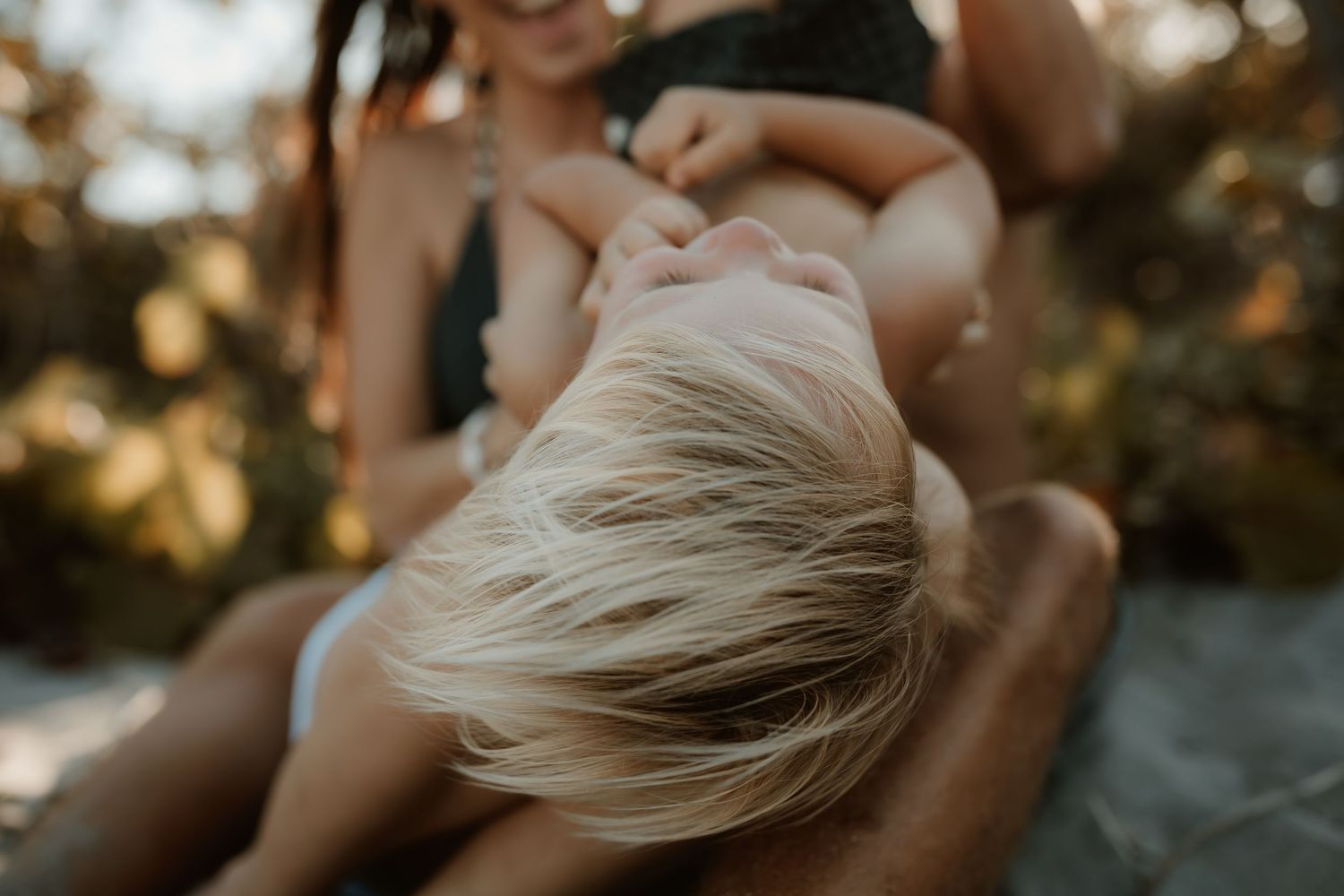 A blonde toddler leans back in a mother's lap outdoors in warm golden light.