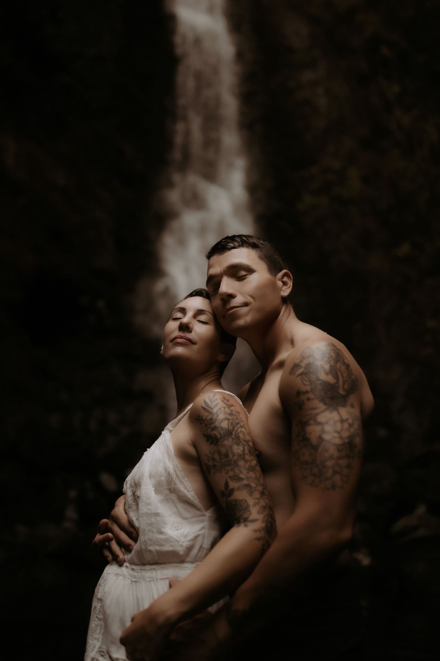 Tattooed couple embracing intimately in front of a misty waterfall, woman in white lace dress, moody dark romantic setting.