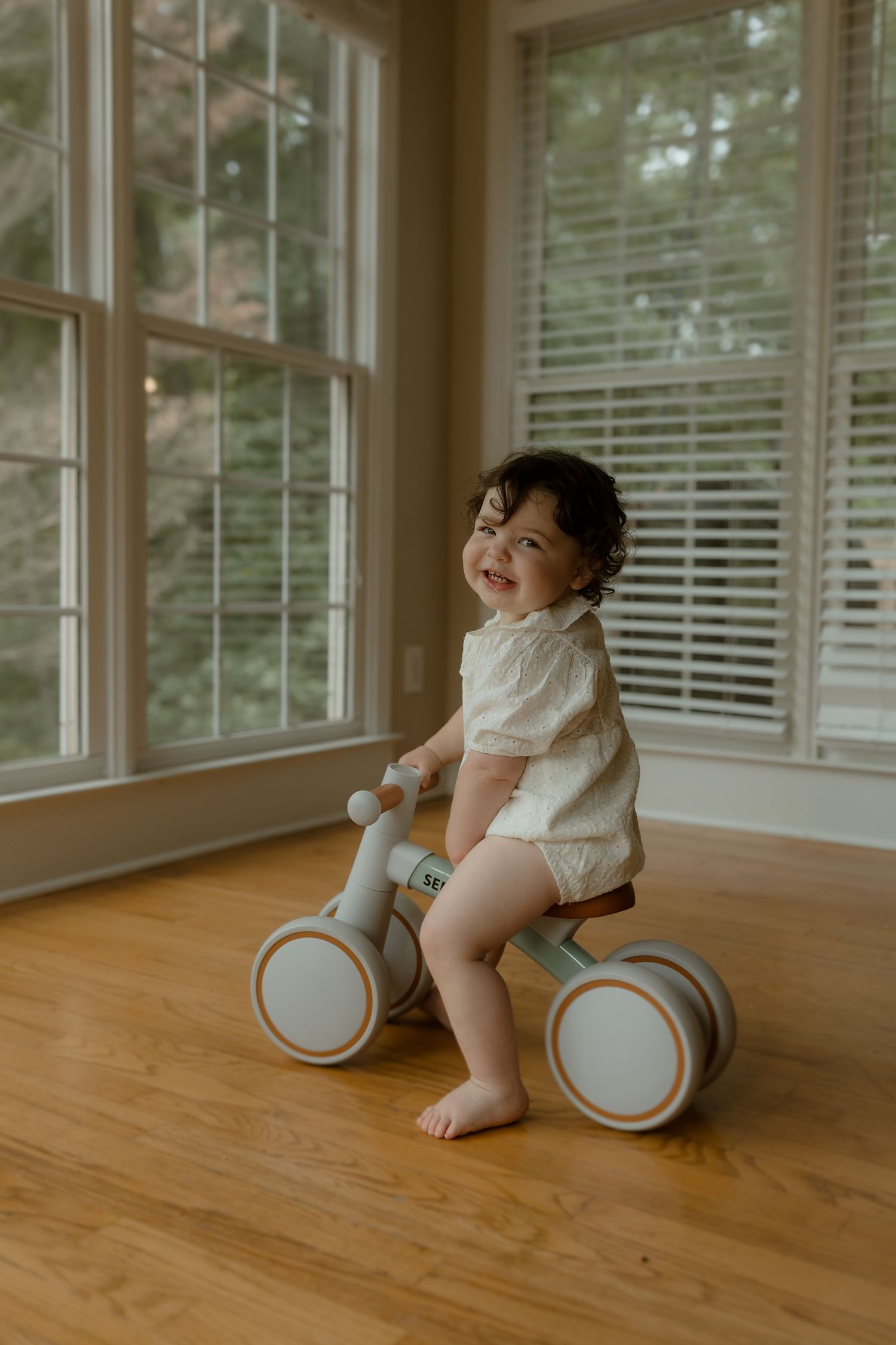Smiling toddler sitting on a gray balance bike toy on hardwood floor near large windows in a bright room.