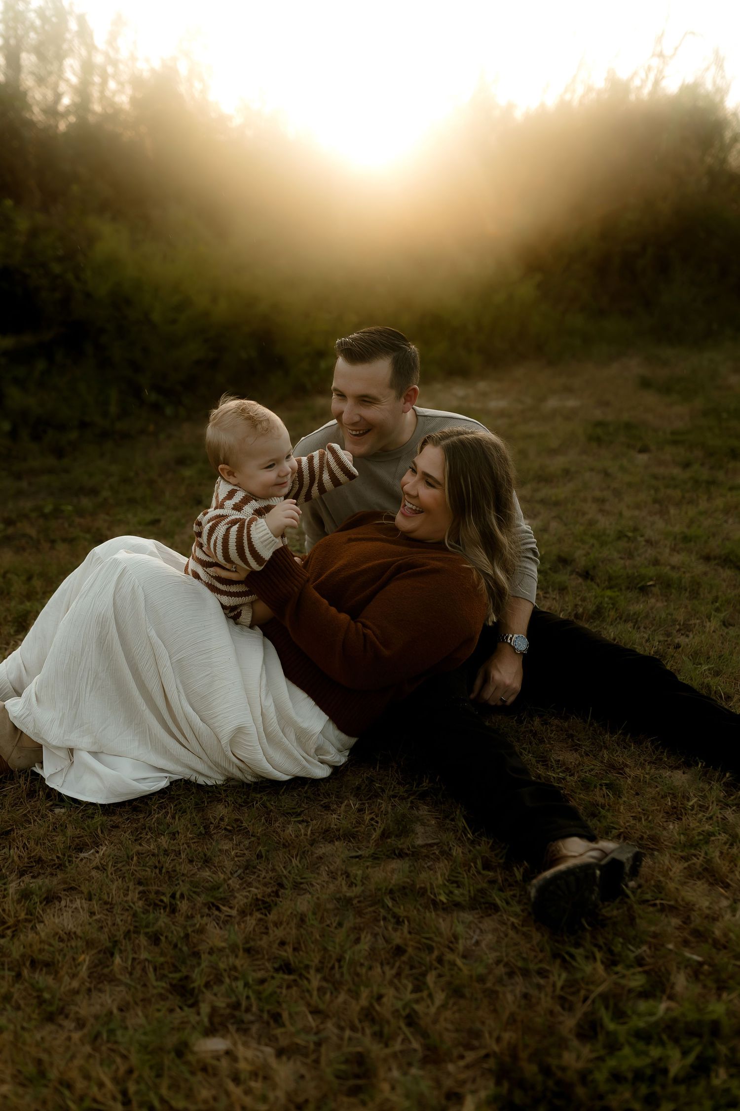 A happy family with a baby sits on grass at golden hour sunset in a lush green outdoor setting.