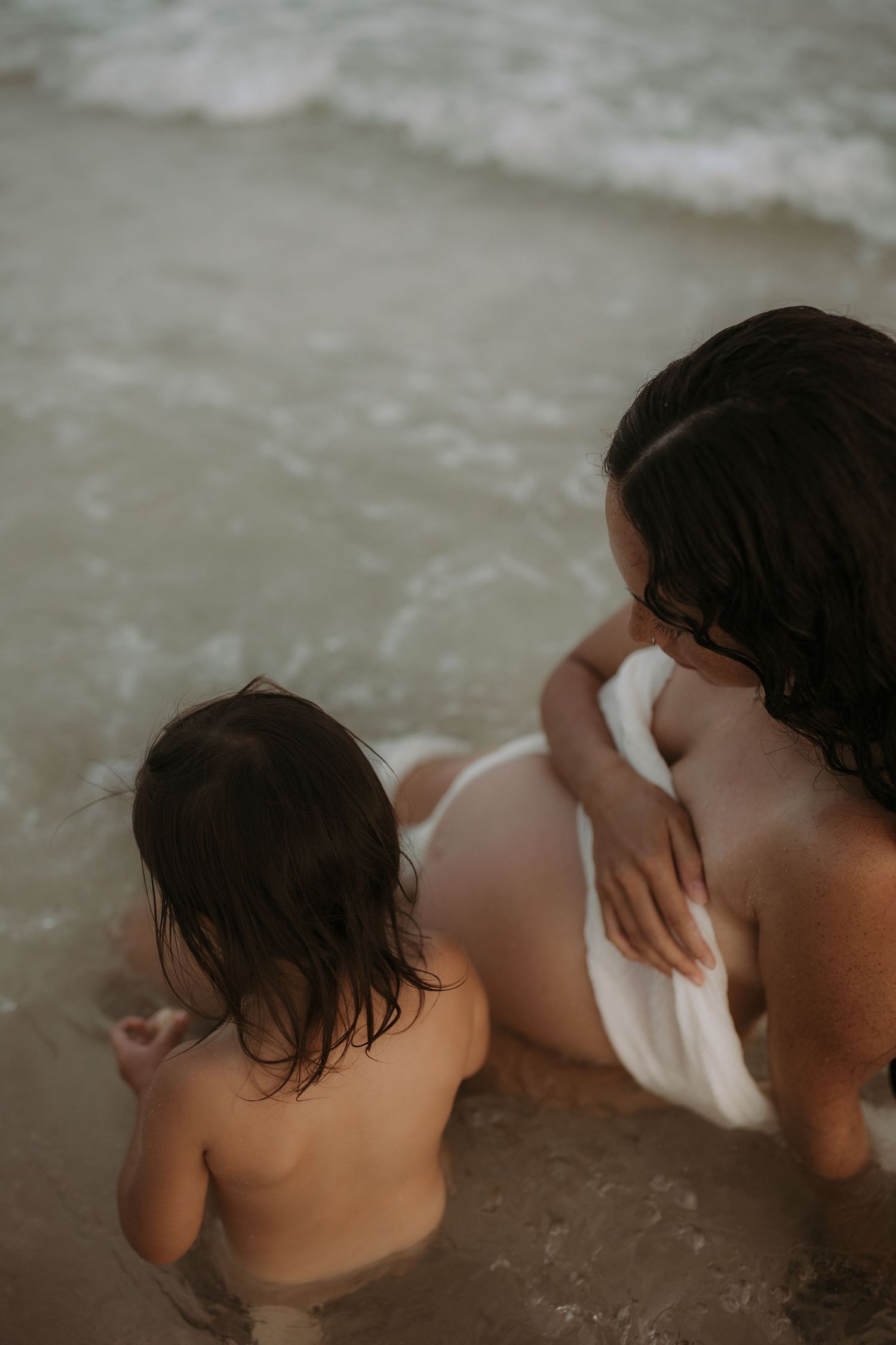 Mother and toddler sitting together in ocean waves at the beach.