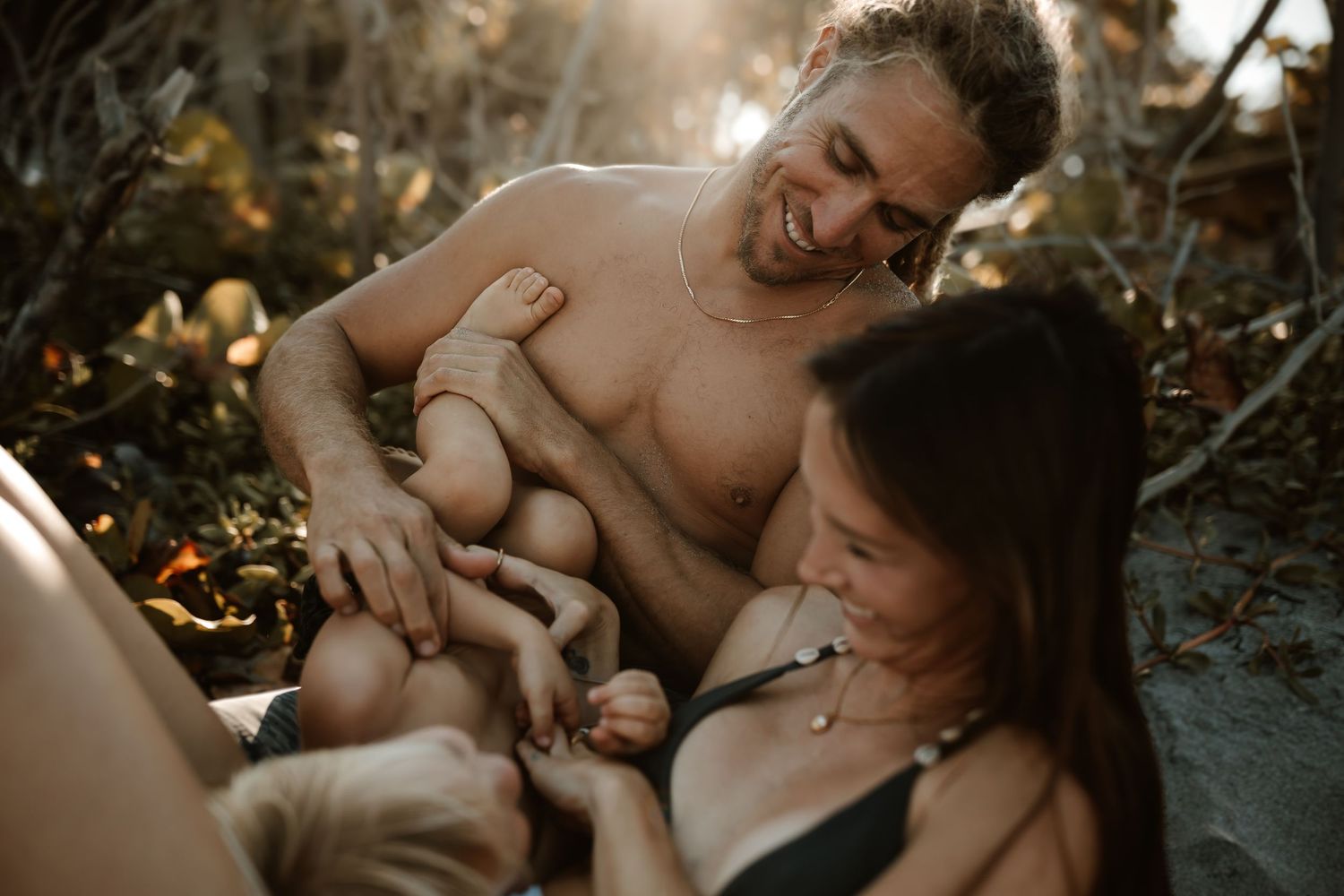 A smiling couple sits outdoors in golden light, tenderly caring for their newborn baby in a natural setting.