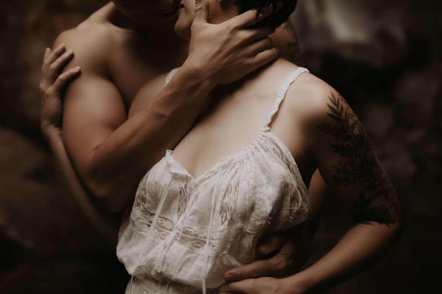 Intimate couple embracing romantically, woman in lace dress with tattoo, moody dark background.