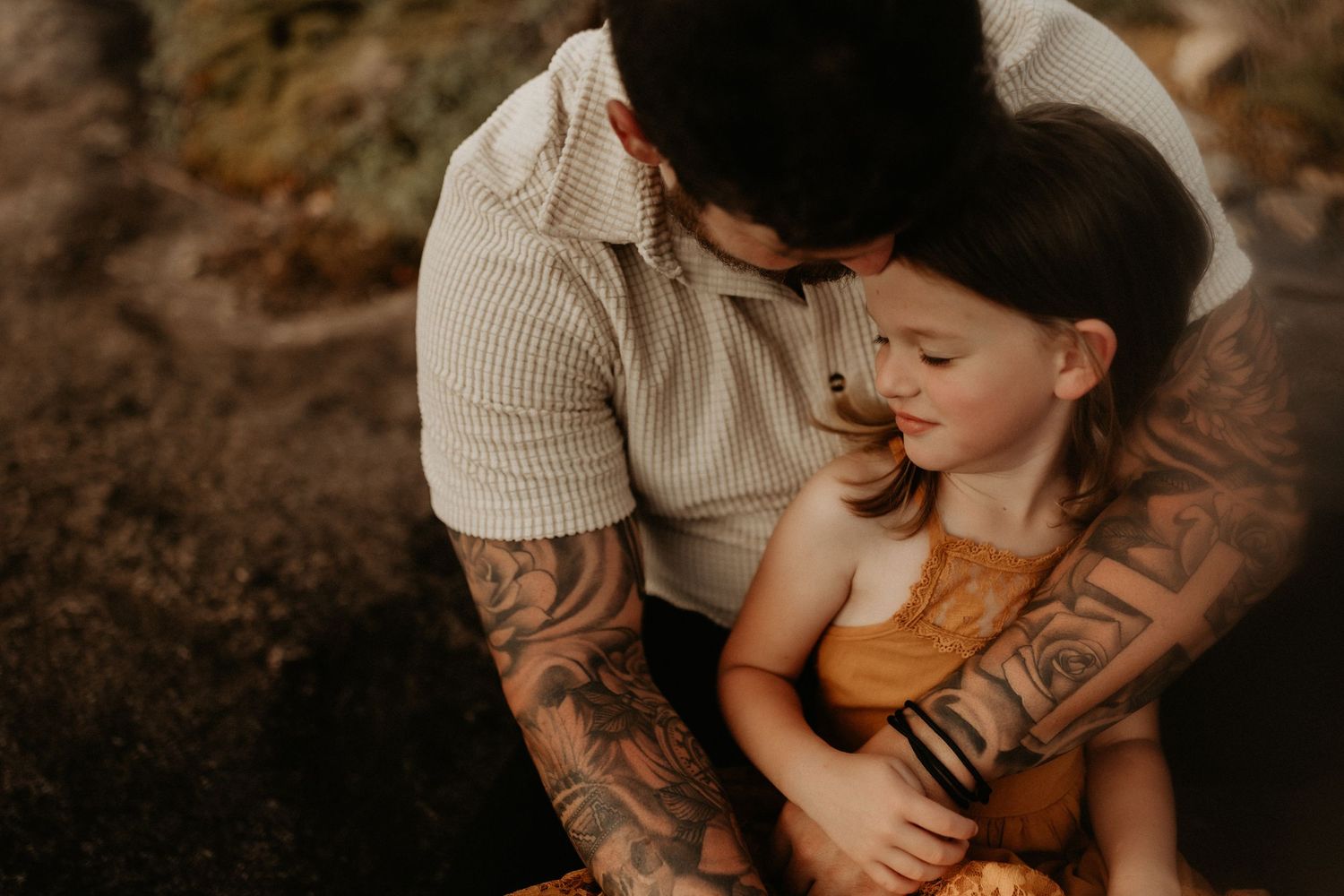 Tattooed father tenderly embraces young daughter in warm golden light outdoor family portrait.