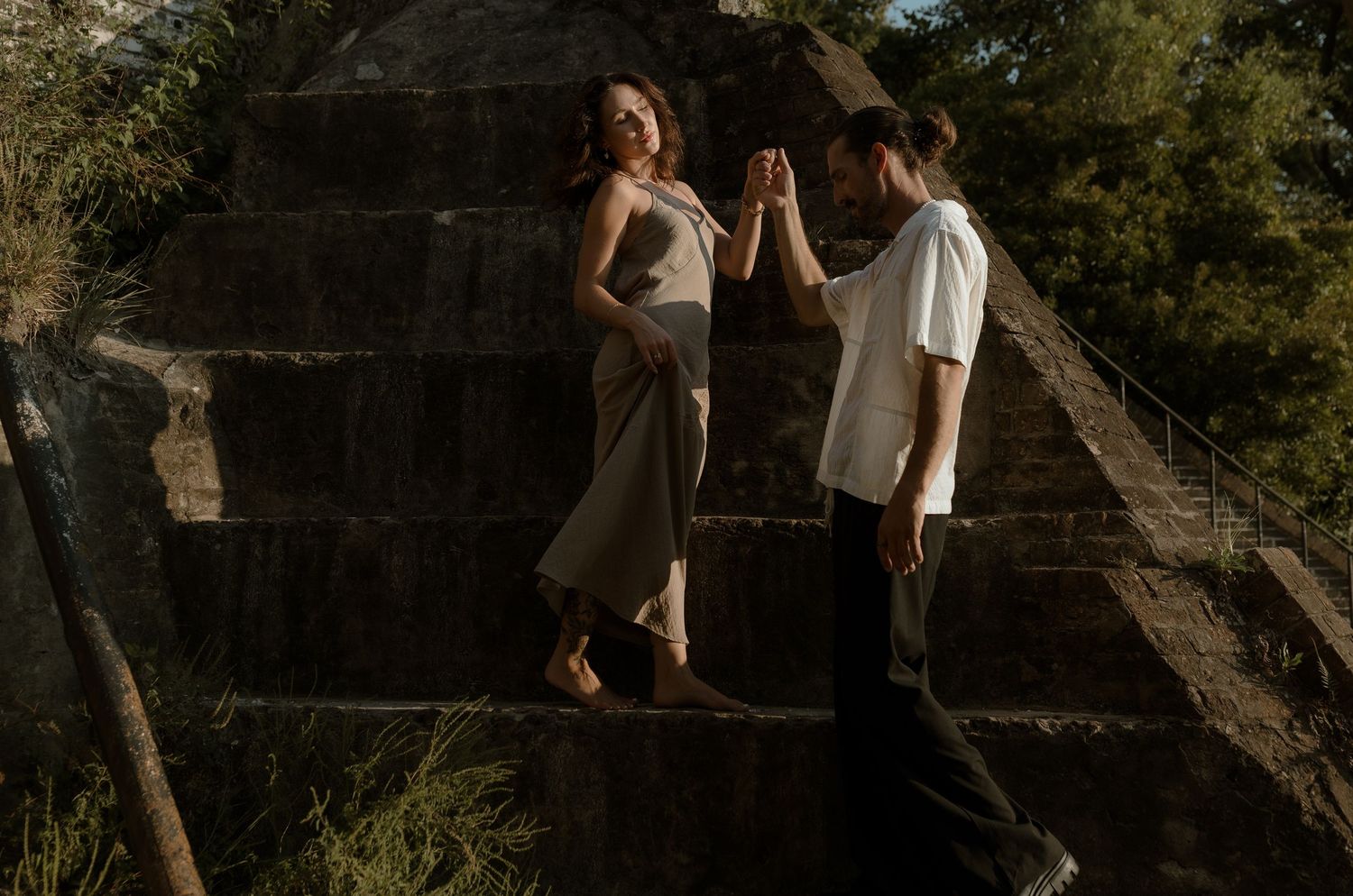 Couple holding hands on stone steps outdoors, woman in gray dress and man in white shirt, surrounded by lush greenery.