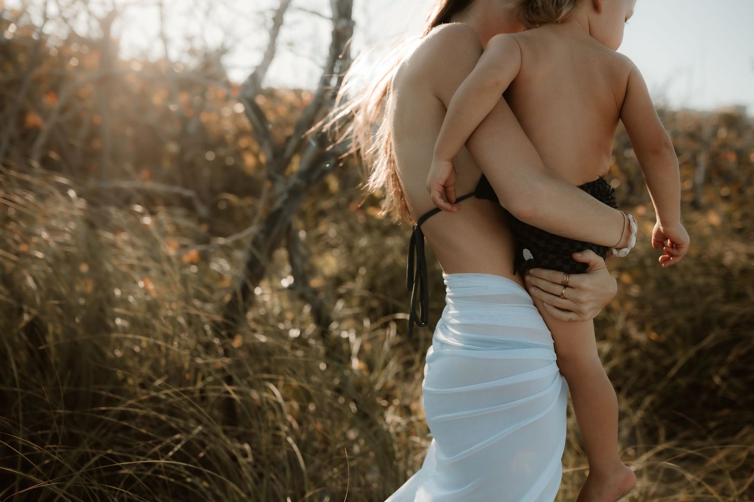 Woman in white skirt holding a young child outdoors in golden sunlight among dry grasses and shrubs.