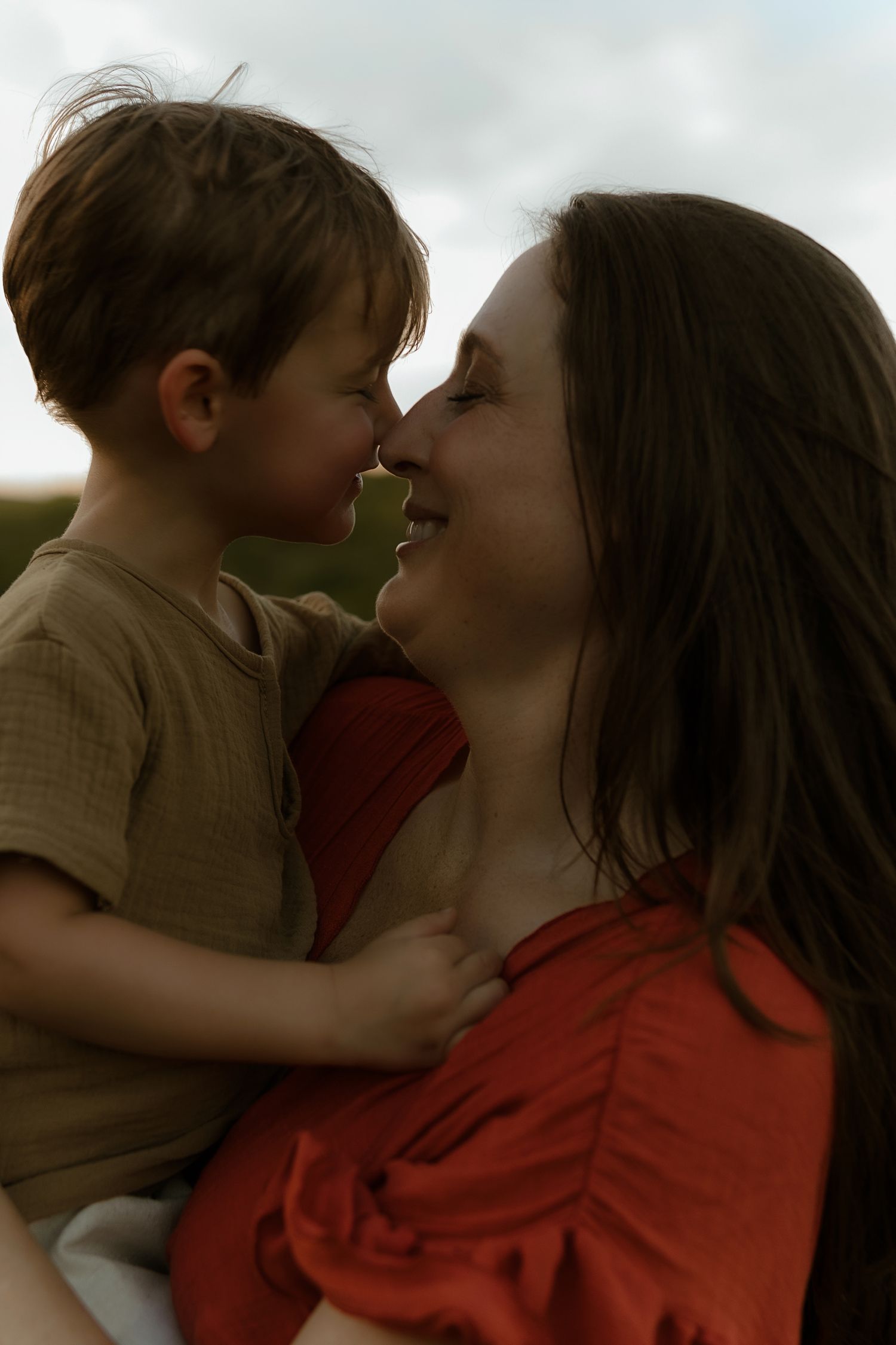 A mother in a red dress tenderly touches noses with her laughing toddler son outdoors.