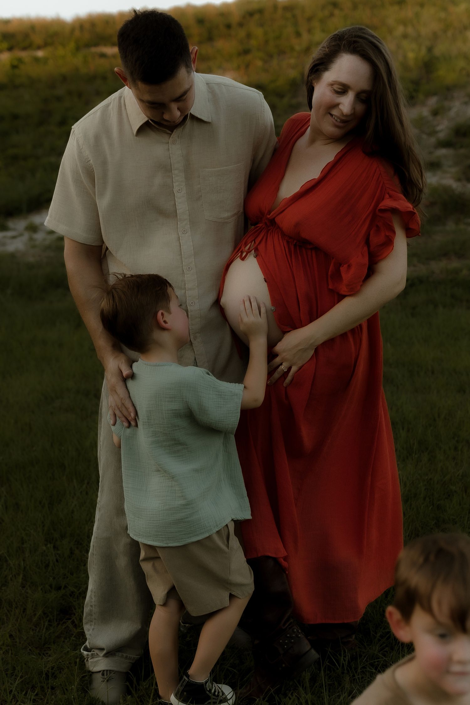A pregnant woman in a red dress with her partner and young child touching her baby bump outdoors.