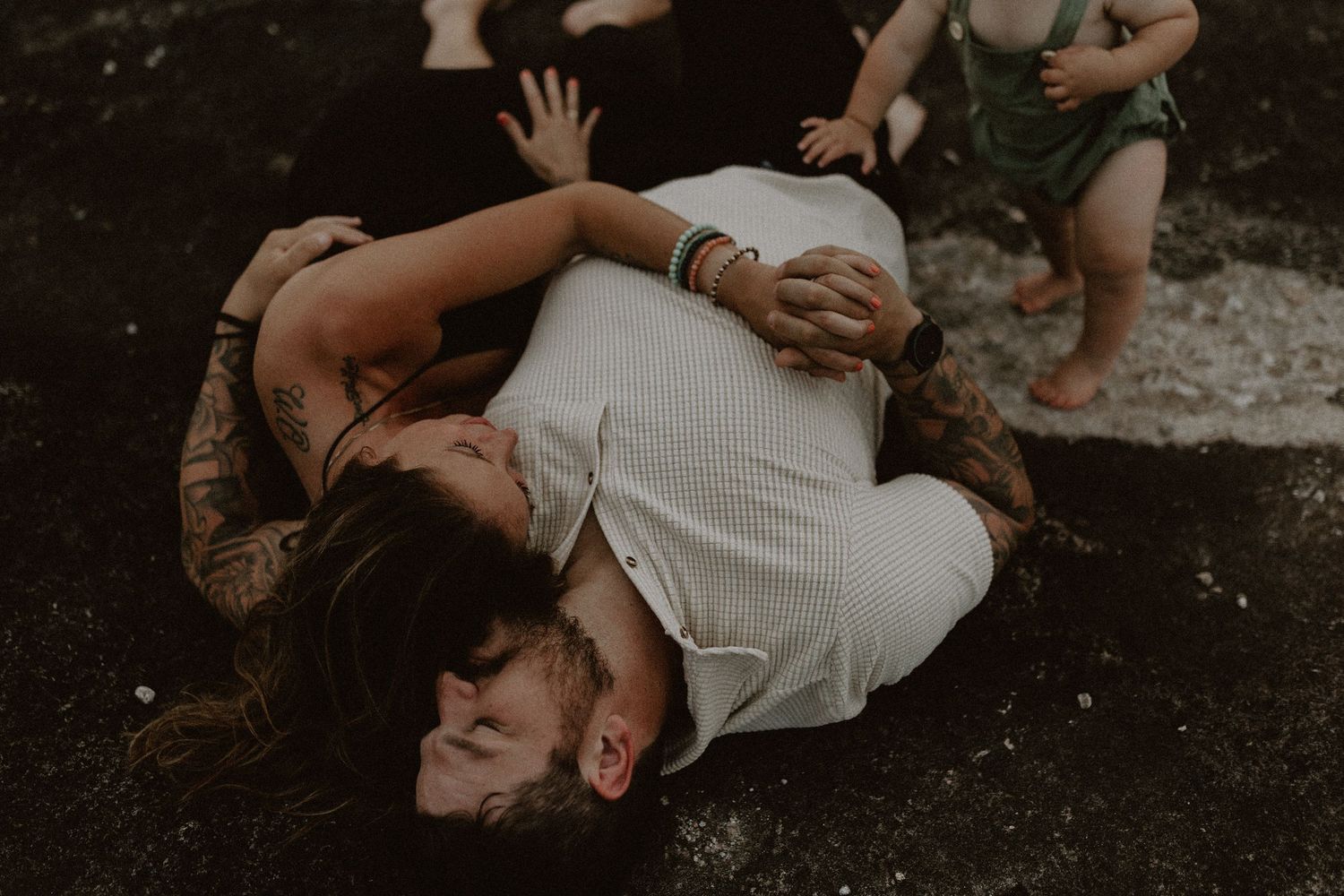 Tattooed couple lying on grass kissing while a toddler plays nearby in a candid family moment.
