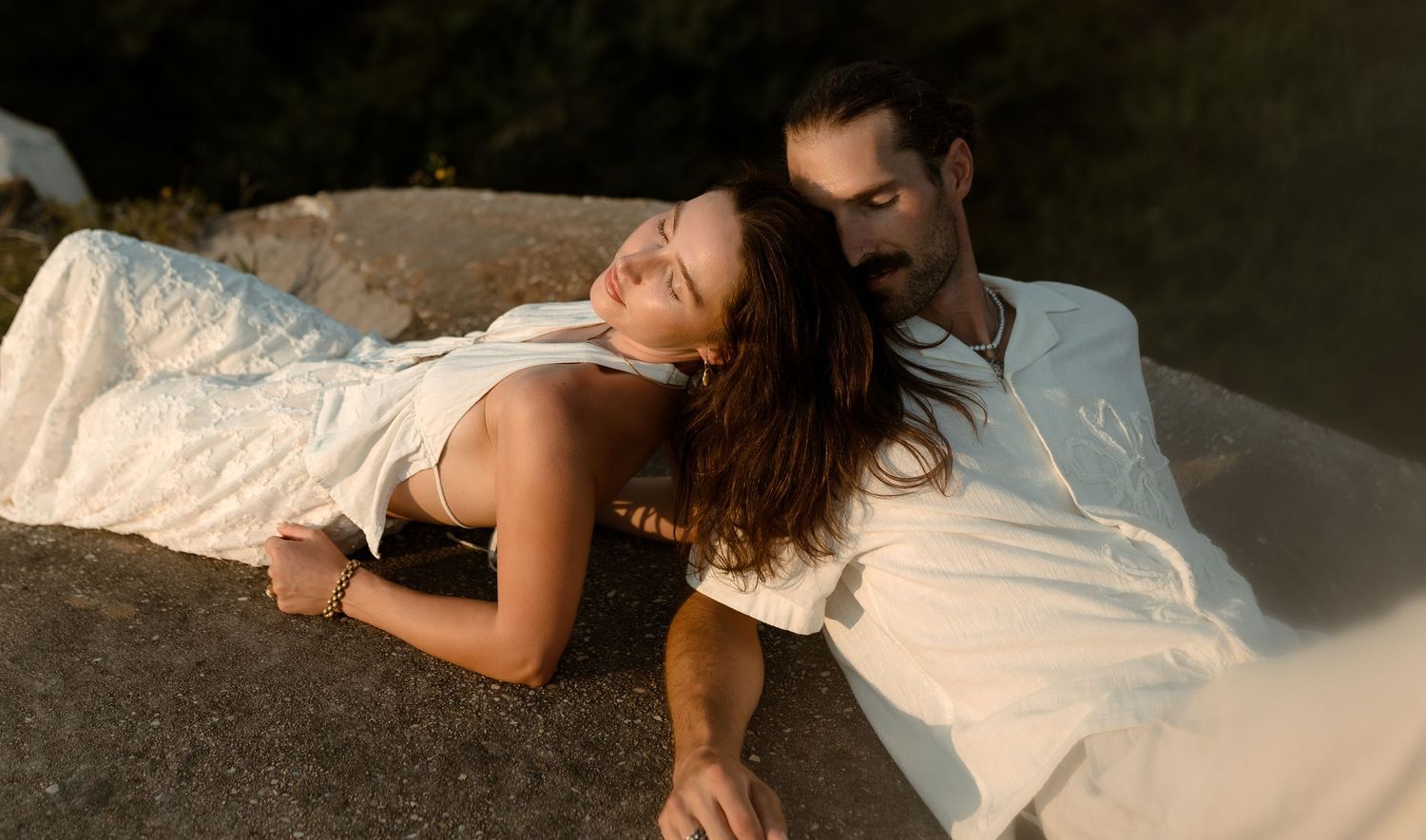 Romantic couple in white outfits resting on a rock outdoors in warm golden sunlight.