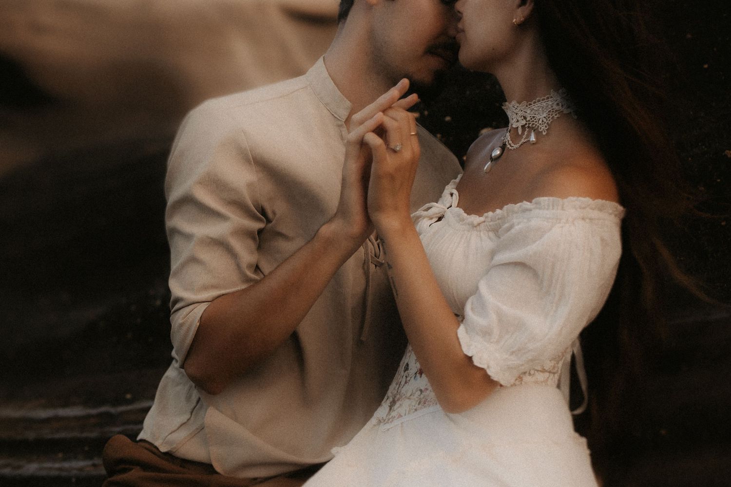 A couple in elegant wedding attire share a tender kiss while holding hands in a romantic outdoor setting.