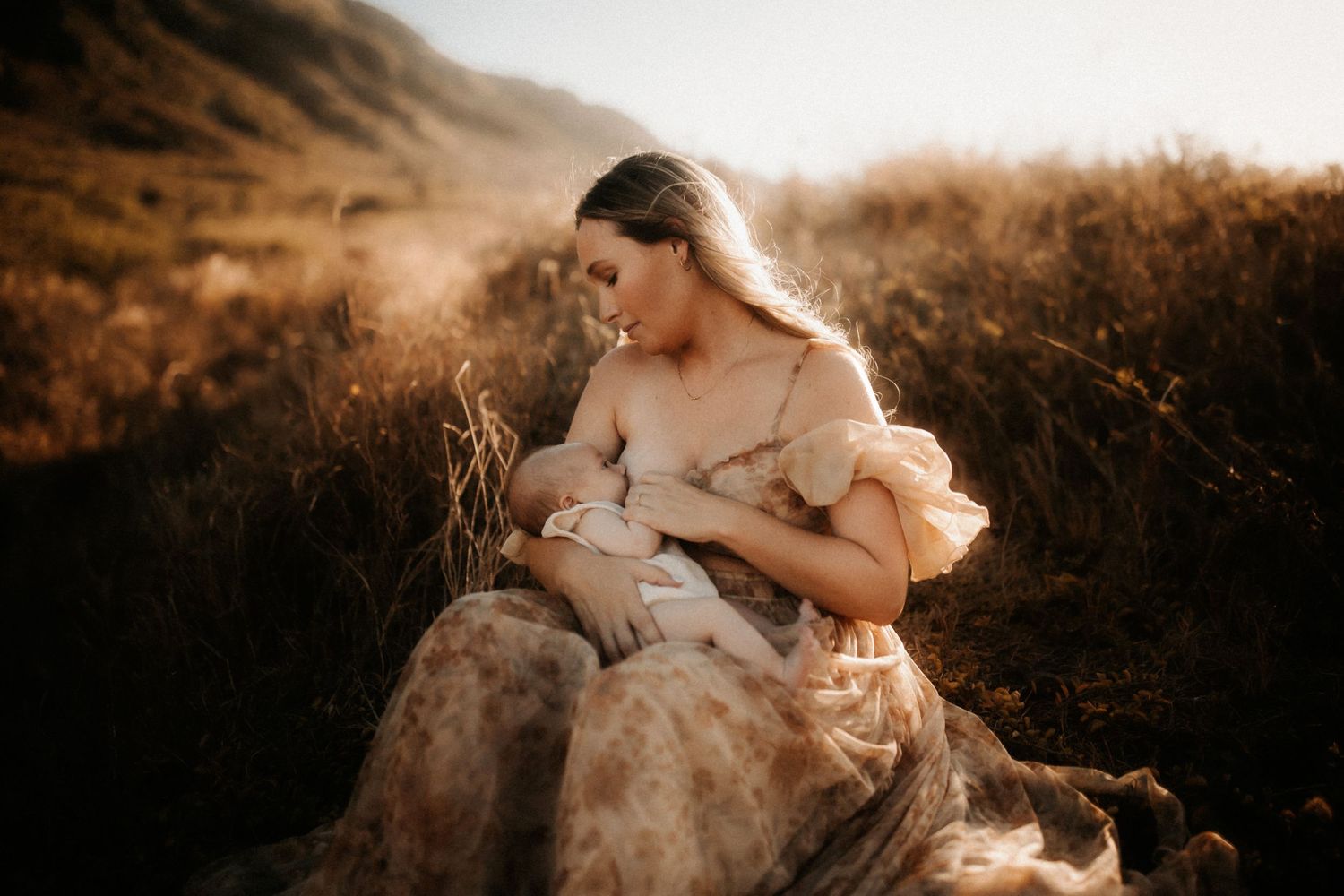 Mother in floral dress breastfeeding baby in golden sunlit field at sunset.