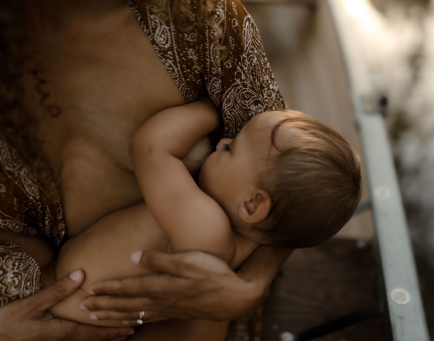 Mother breastfeeding infant in warm natural light wearing brown floral dress outdoors.