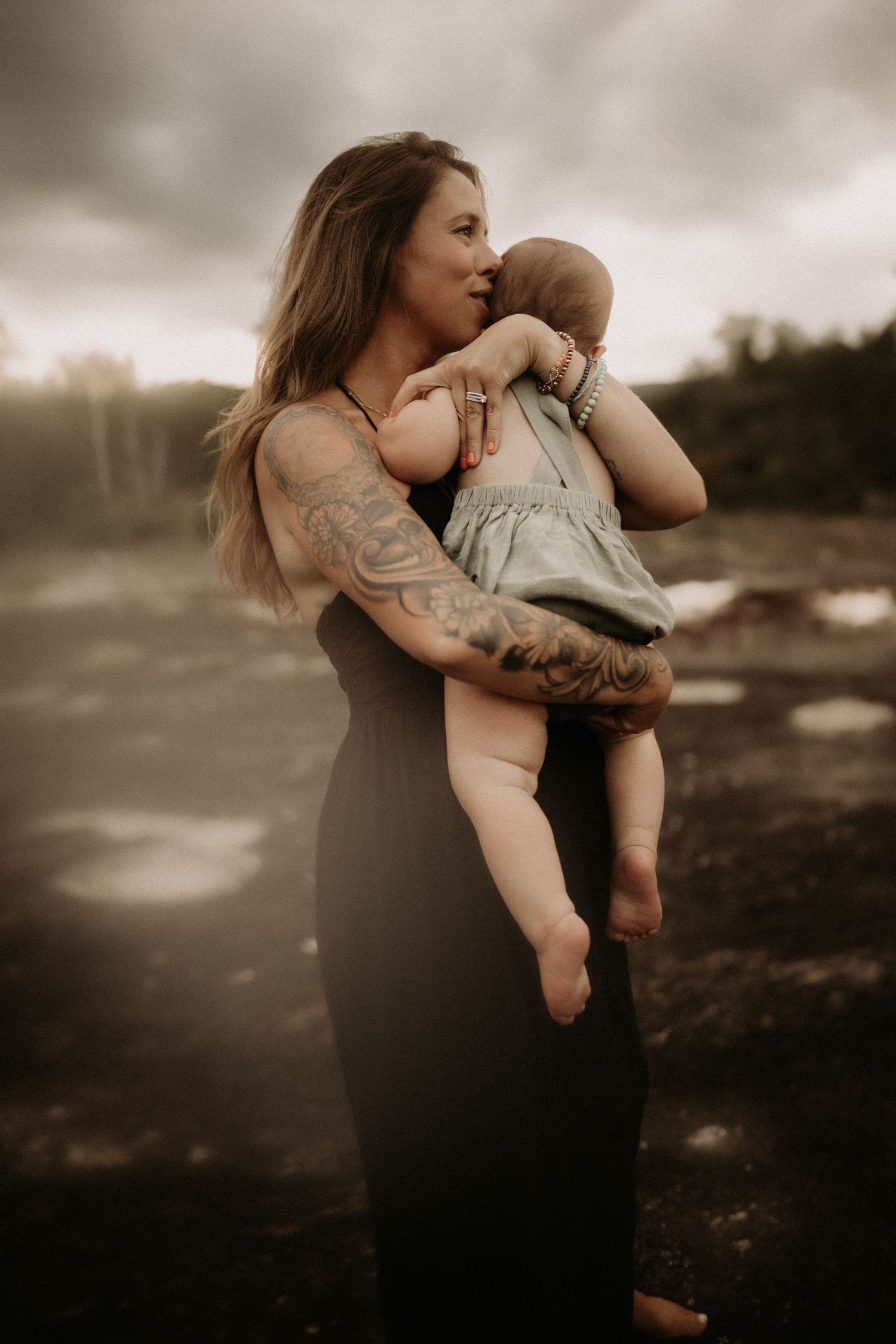Tattooed mother in black dress tenderly holding her baby under dramatic cloudy skies outdoors.