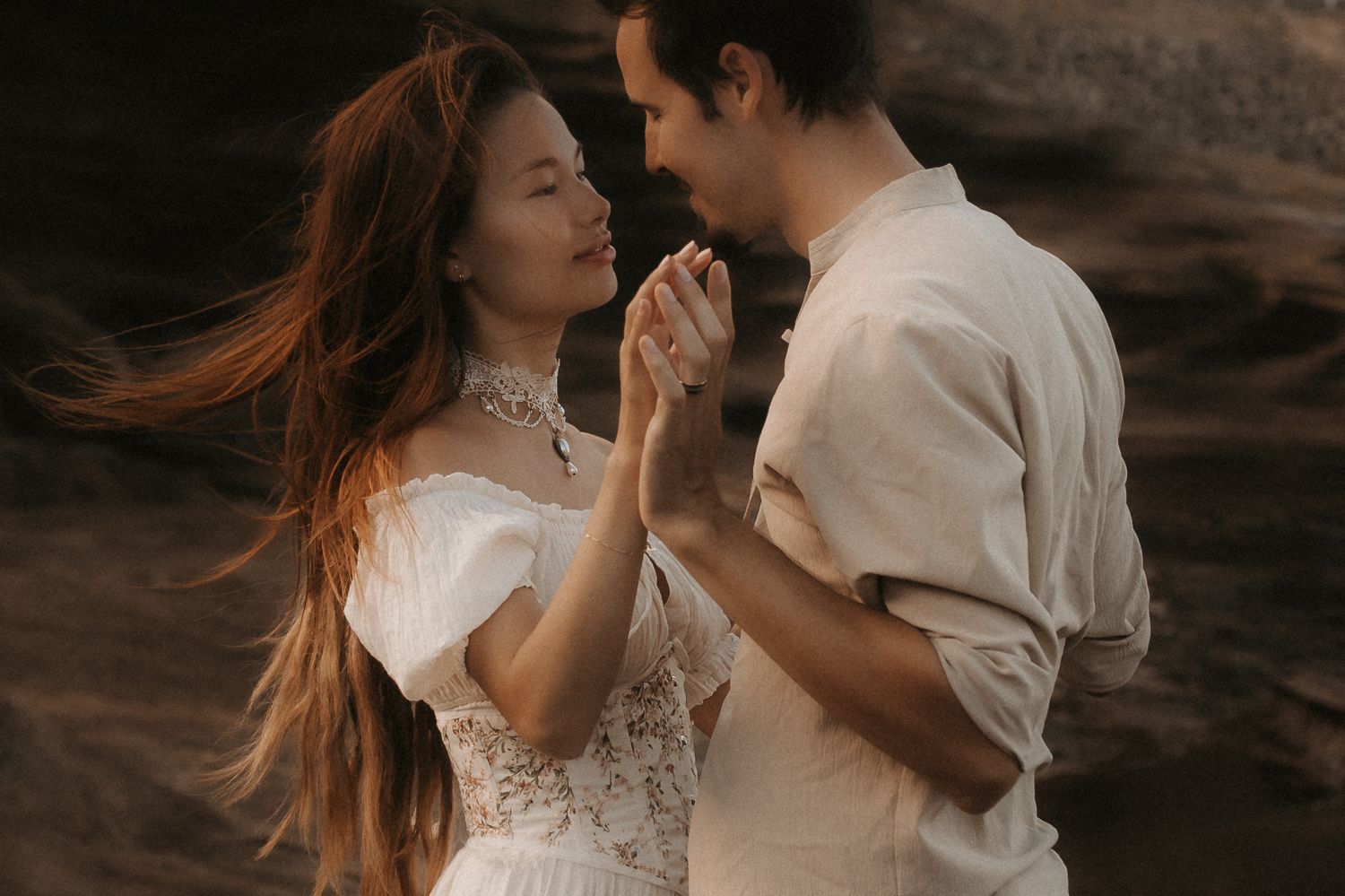 Romantic couple in white attire holding hands and gazing at each other outdoors near rocky cliffs.