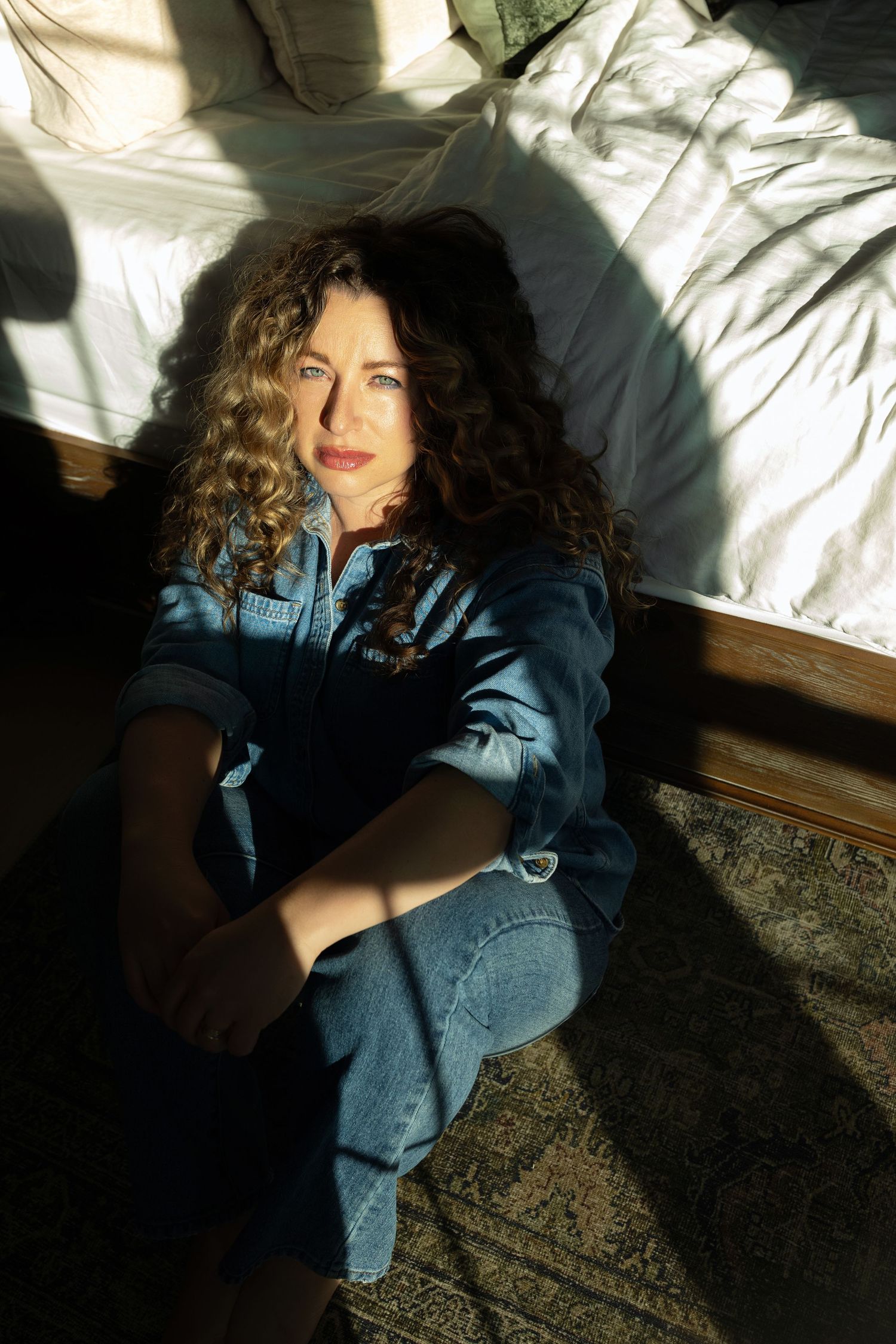 Woman with curly hair in denim outfit sits on floor leaning against bed in sunlight.