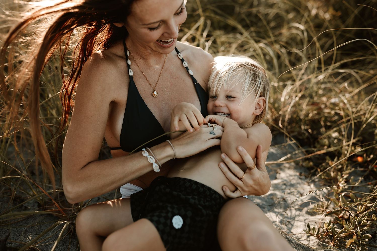 A mother laughs with her toddler on a sunny beach, both wearing black swimwear amid golden grass.