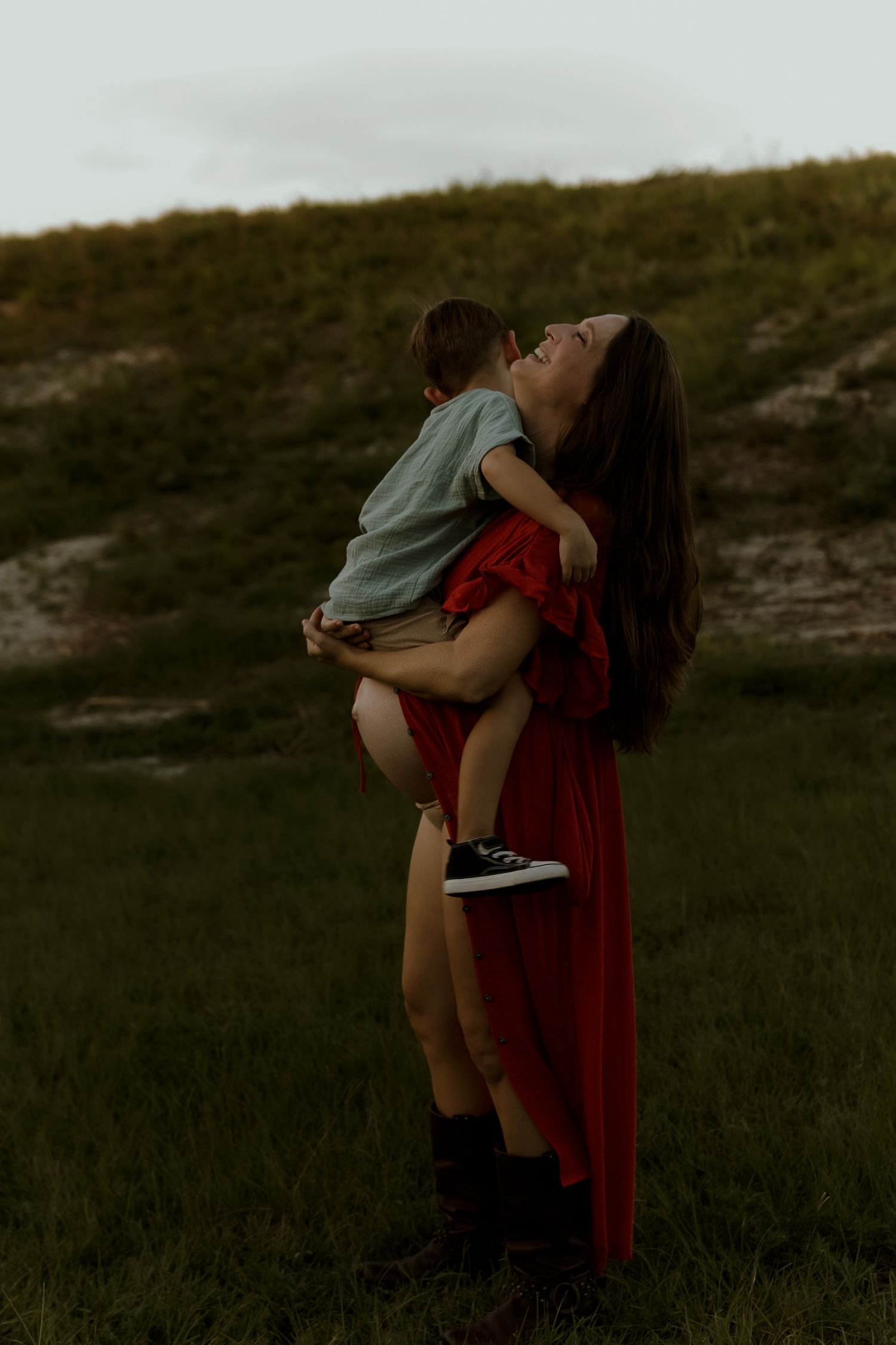 A woman in a red dress holds a toddler outdoors on a grassy hill at dusk, both laughing joyfully.